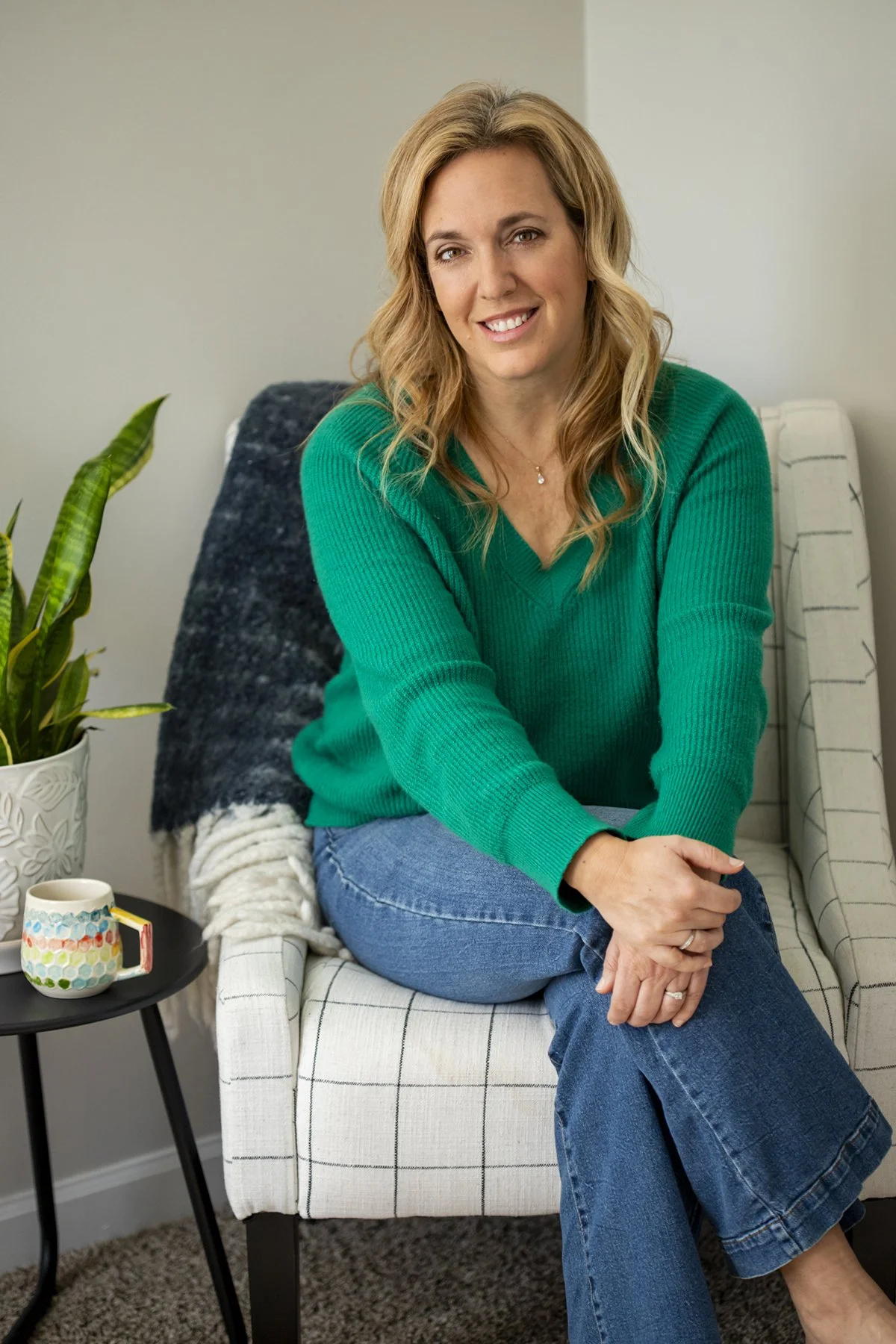 A woman with wavy blonde hair smiling, seated on a white and black checkered armchair, wearing a green sweater and blue jeans, with her hands resting on her knee. There is a houseplant on a small black table next to her, along with a colorful mug and a white blanket with dark grey pattern on the chair.