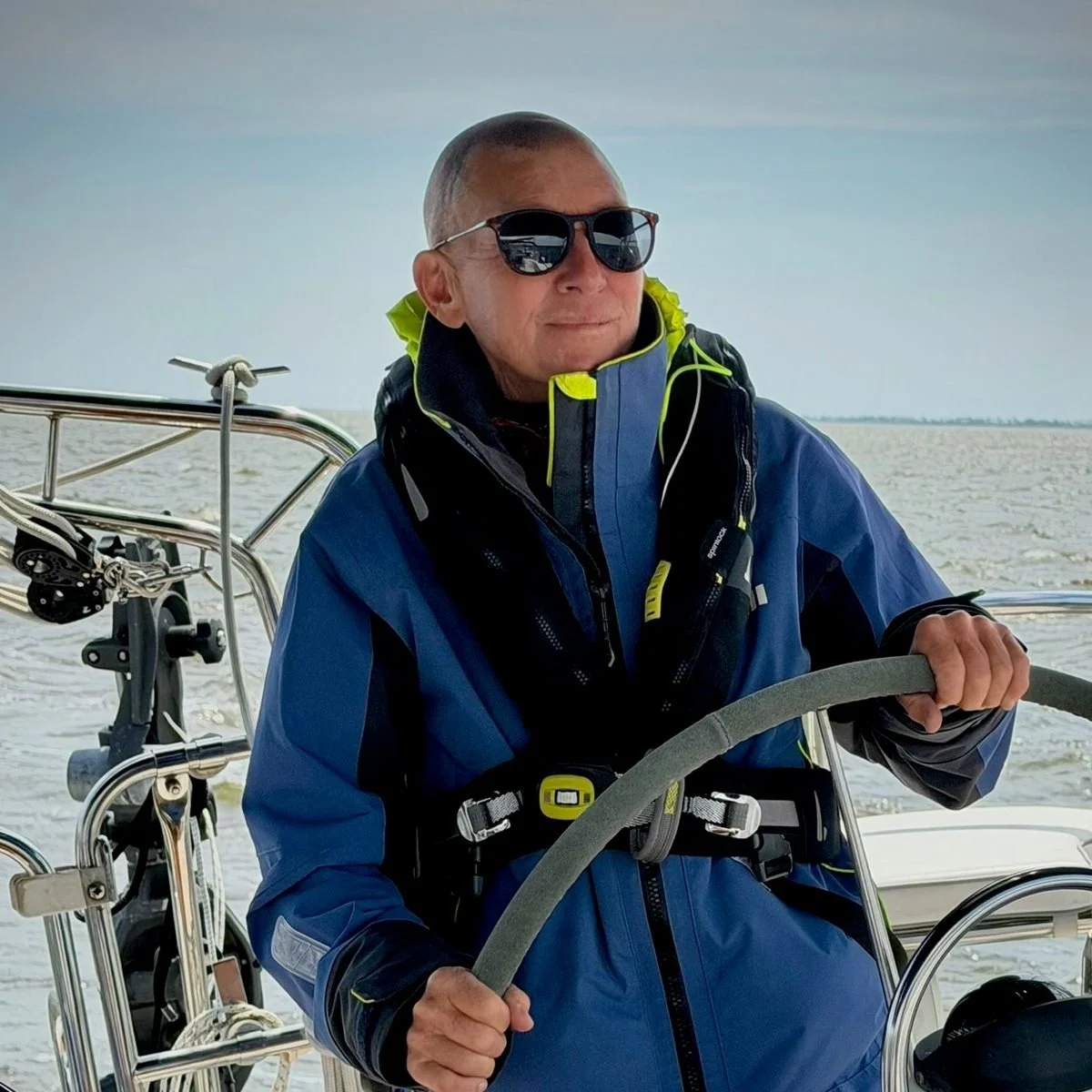 Captain e2 Brewster at the helm of a sailboat in a blue jacket and life vest, two hands on the wheel.