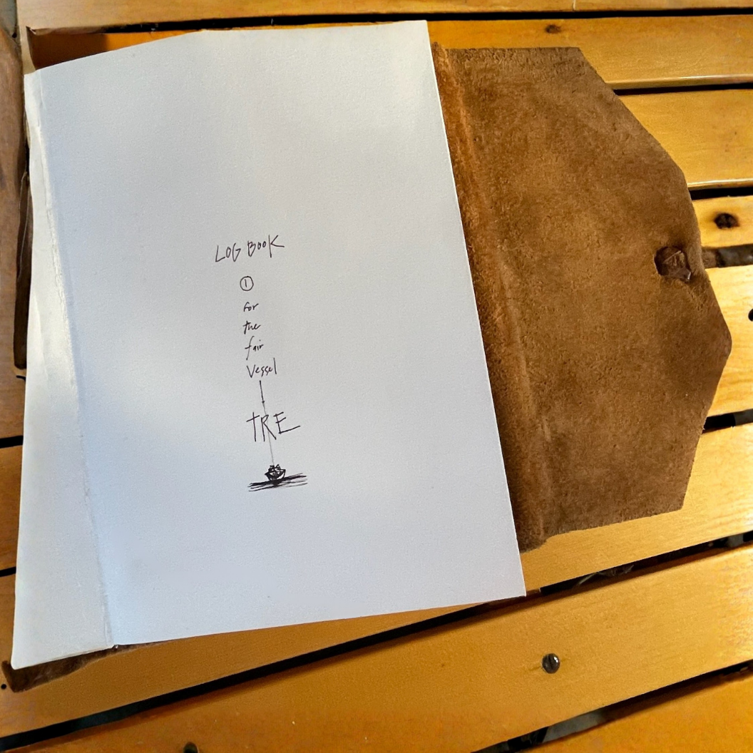 Open handmade leather notebook with a handwritten title 'Log Book' and the subtitle 'for the fair vessel TRE' with a small doodle of a boat at the bottom, resting on a watery surface.