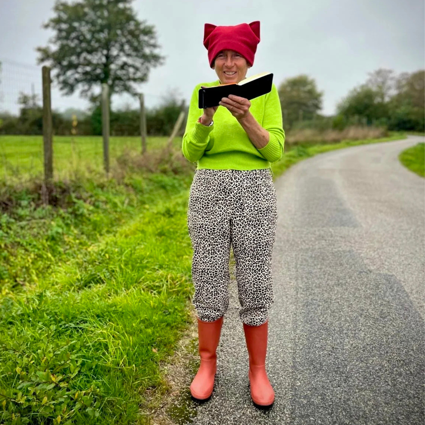 Captain e2 Brewster standing in the road, sketching in their sketchbook — cat-ear hat, lime green sweater, leopard print pants, red boots.