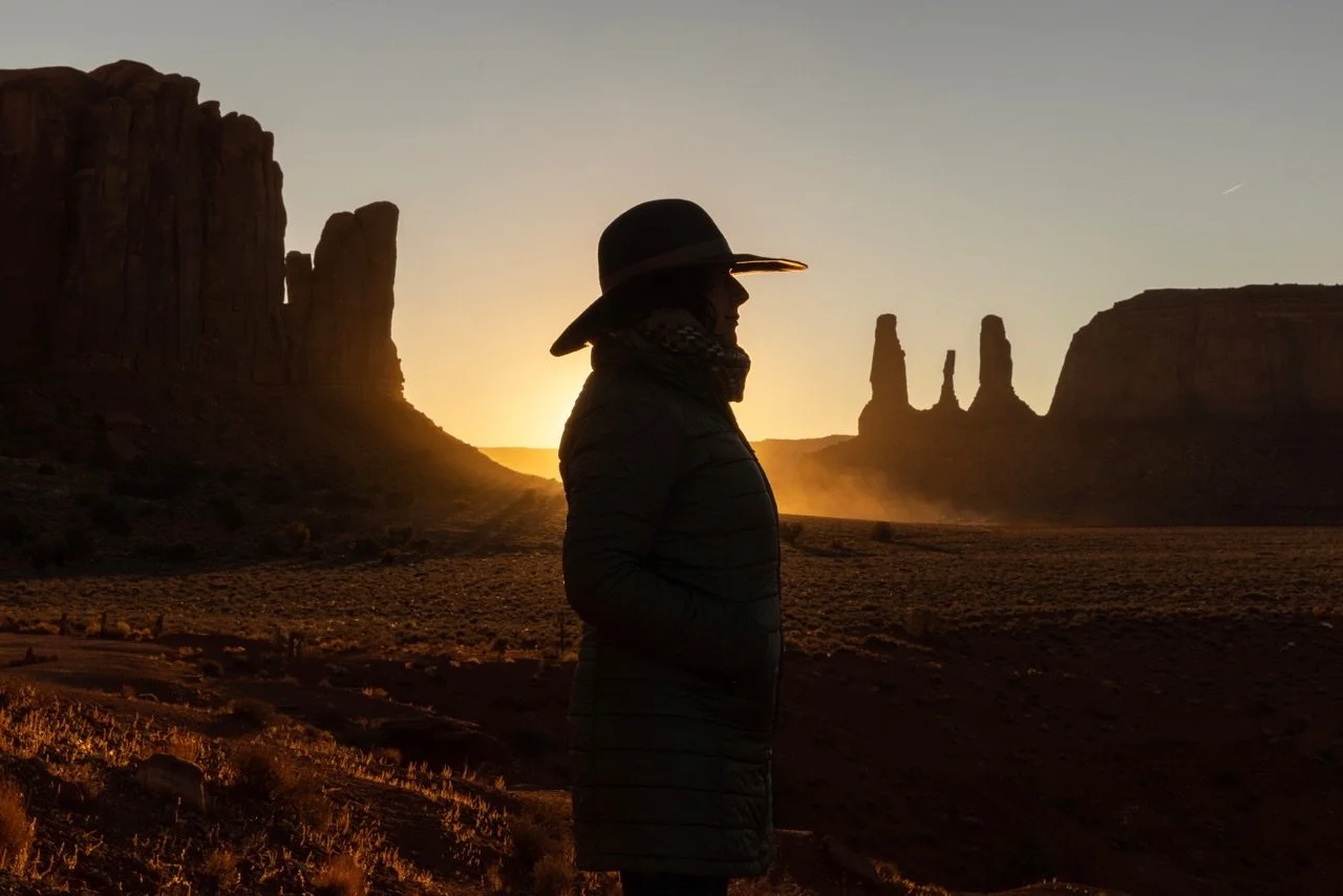 Amy Lee in profile, wearing a hat, in a desert landscape with large rock formations at sunset.