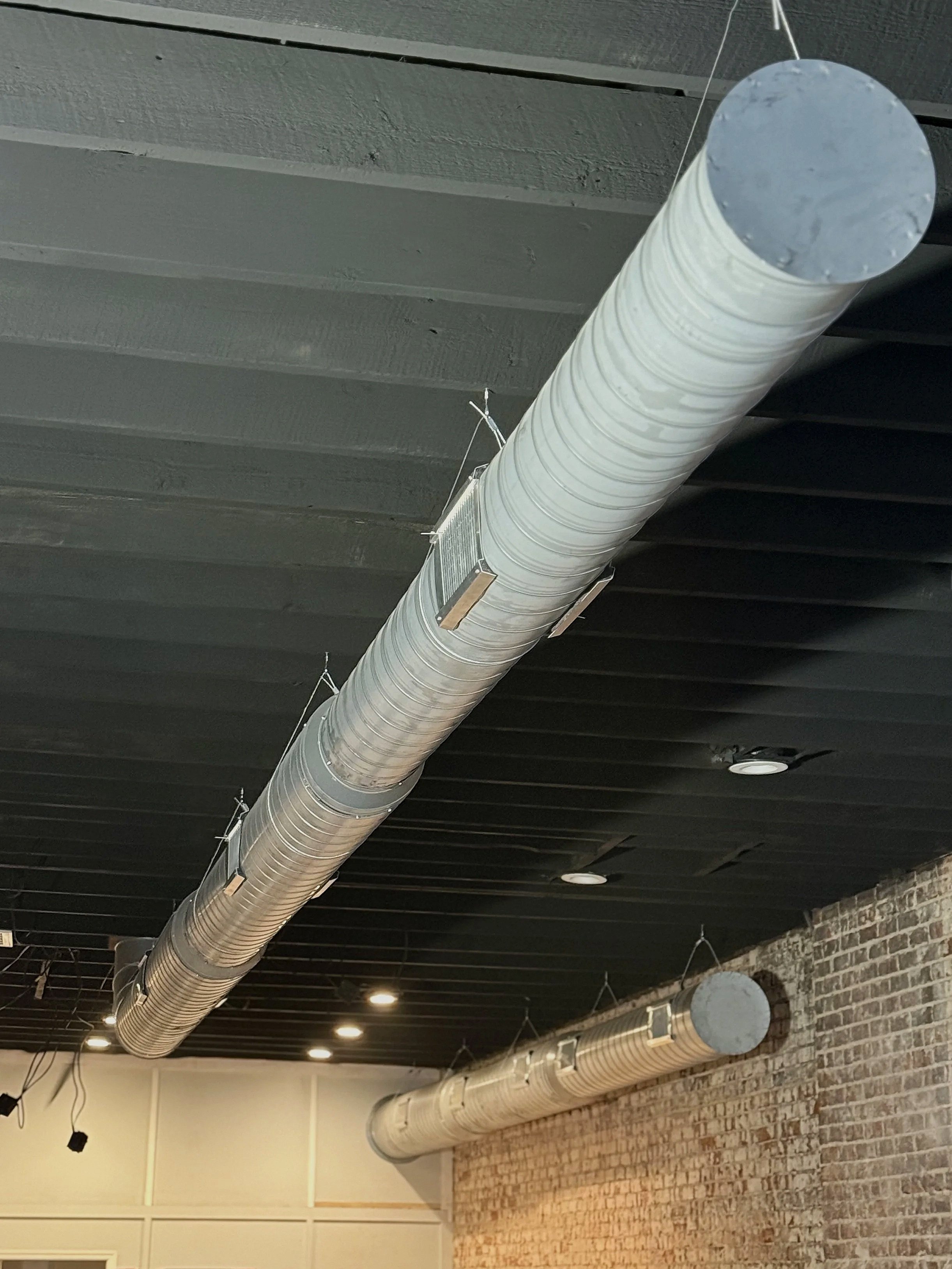 Two large silver industrial air ducts suspended from a black ceiling in a room with brick and white walls.