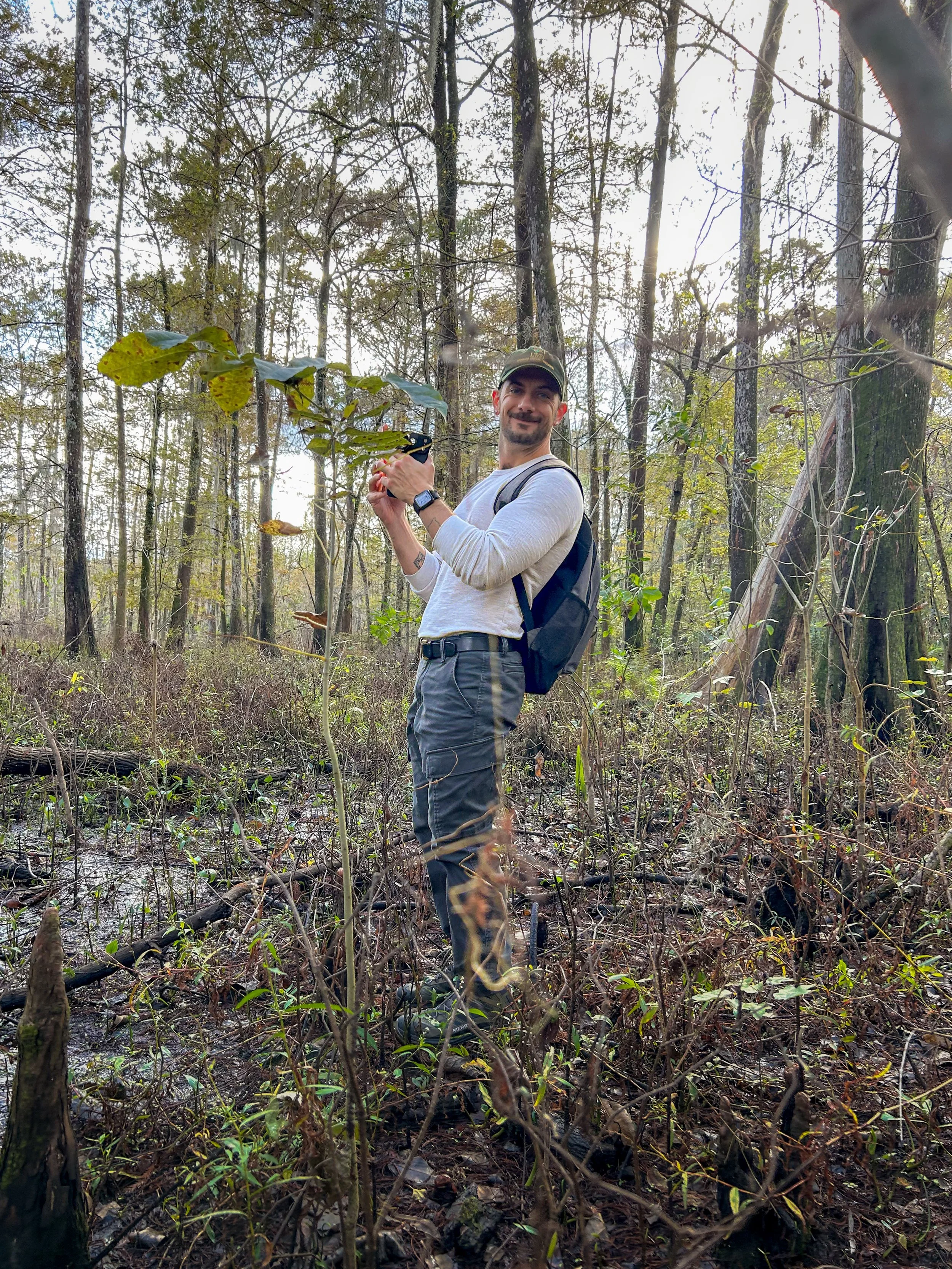 A man with a backpack standing in a swamp, holding a camera and smiling at the camera.