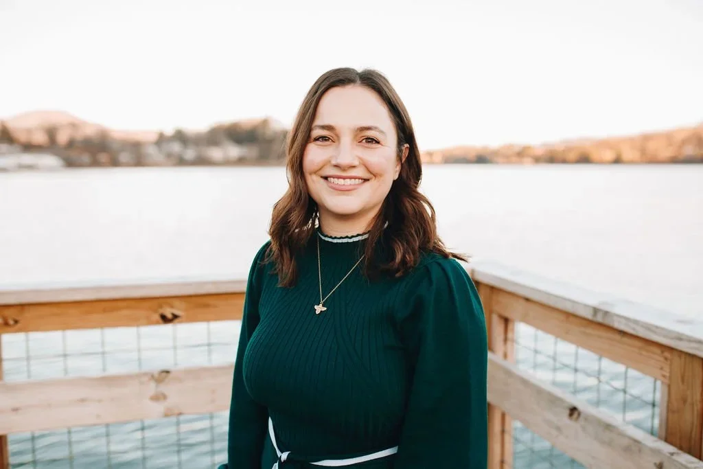 A woman outdoors near a body of water, smiling at the camera, with a wooden railing behind her and a blurred landscape in the background.