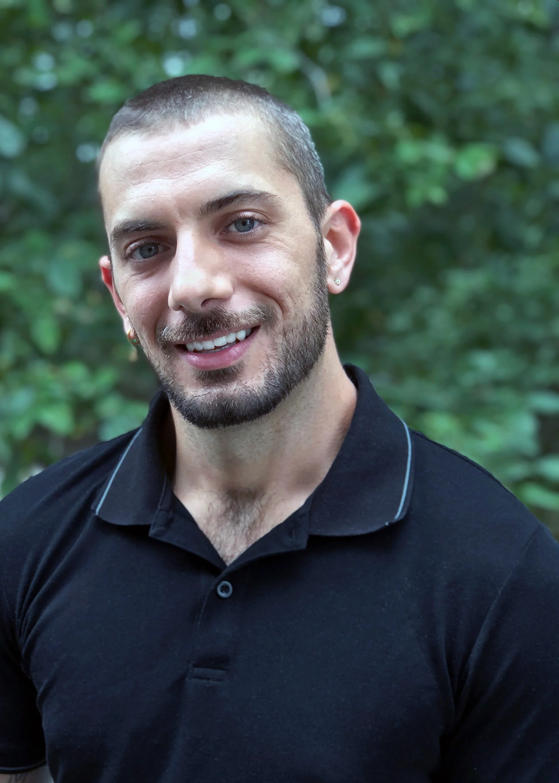 A young man with short brown hair, blue eyes, and a light beard, smiling outdoors with green foliage in the background, wearing a black polo shirt.