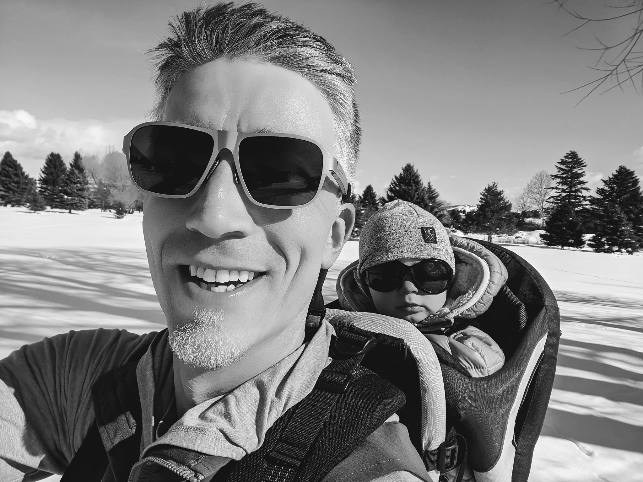 An adult man wearing sunglasses and smiling, carrying a child in a backpack carrier, both dressed warmly in winter clothing, outdoors in a snowy landscape with trees in the background.