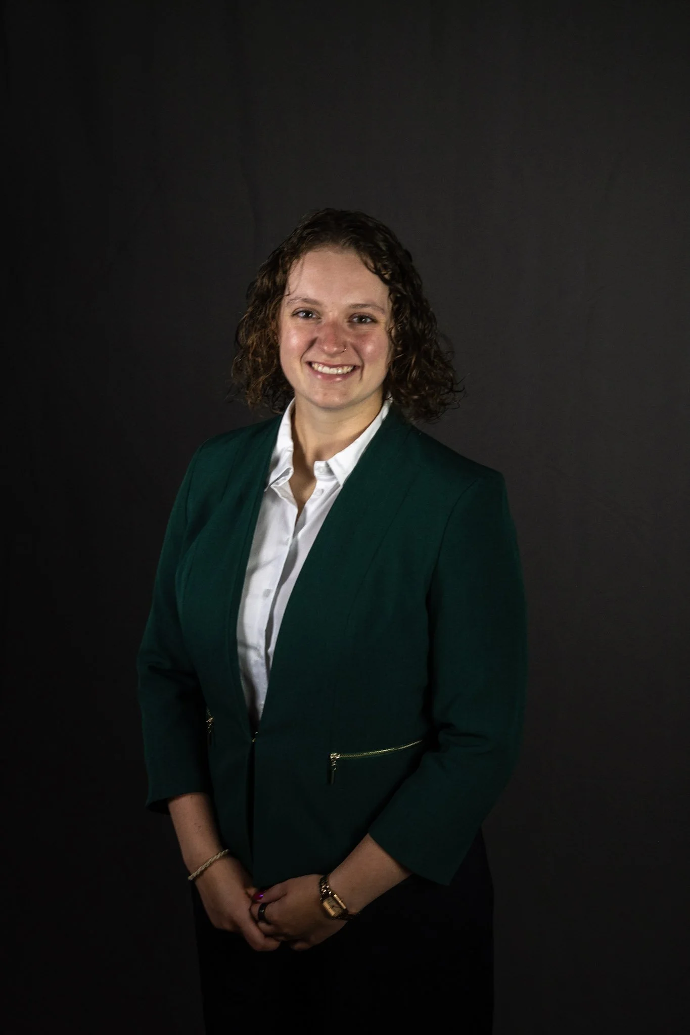 A woman with curly brown hair wearing a dark green blazer and white shirt, standing against a black background, smiling at the camera.