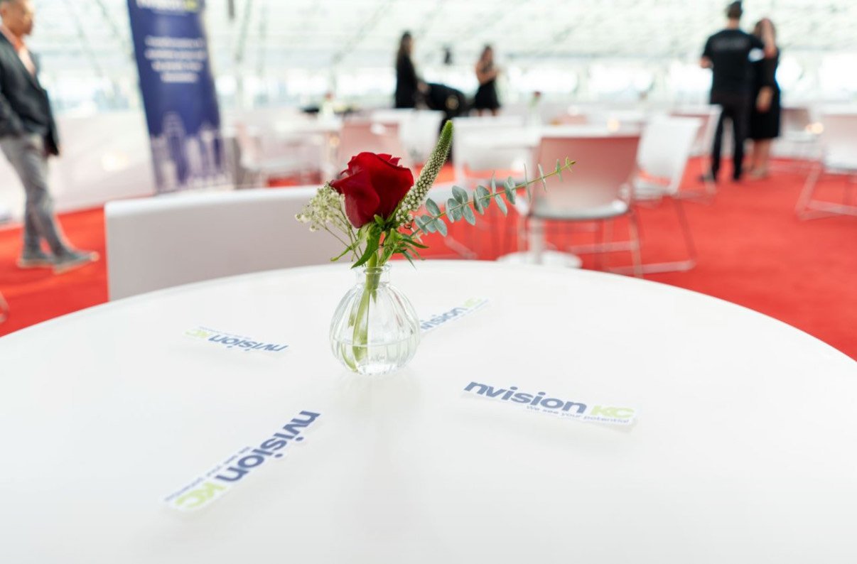 A small glass vase with a red rose, some white flowers, and eucalyptus leaves on a white table at an event, with blurred people and chairs in the background.