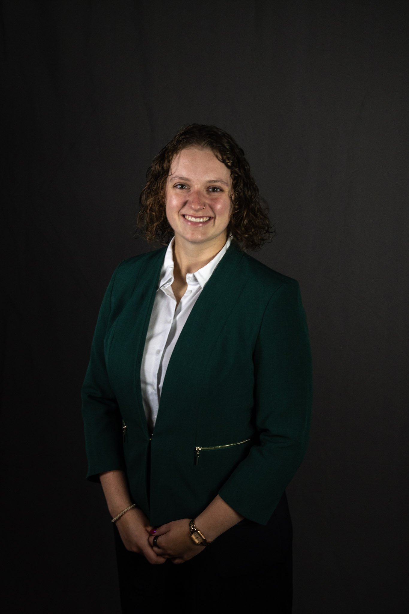 A woman with curly brown hair wearing a dark green blazer over a white shirt, standing against a dark background, smiling at the camera.