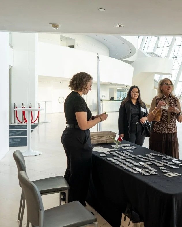 Three women stand around a black table with name tags and small items, in a modern, bright lobby with white walls and large windows.