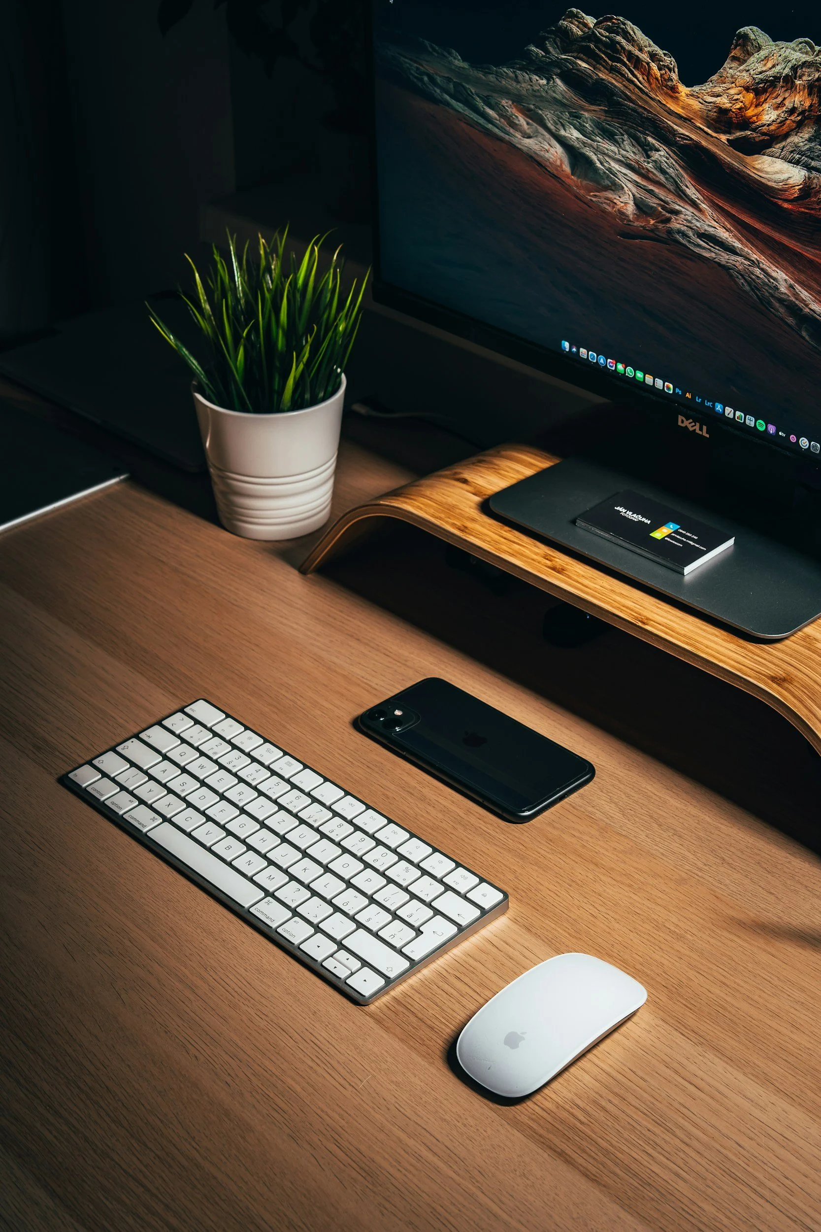 A neat workspace with a wooden desk holding an Apple Magic Keyboard and Magic Mouse, a black iPhone, a potted plant, and a Dell monitor displaying a mountain landscape wallpaper. A laptop with a wooden stand and a closed notebook are also on the desk.