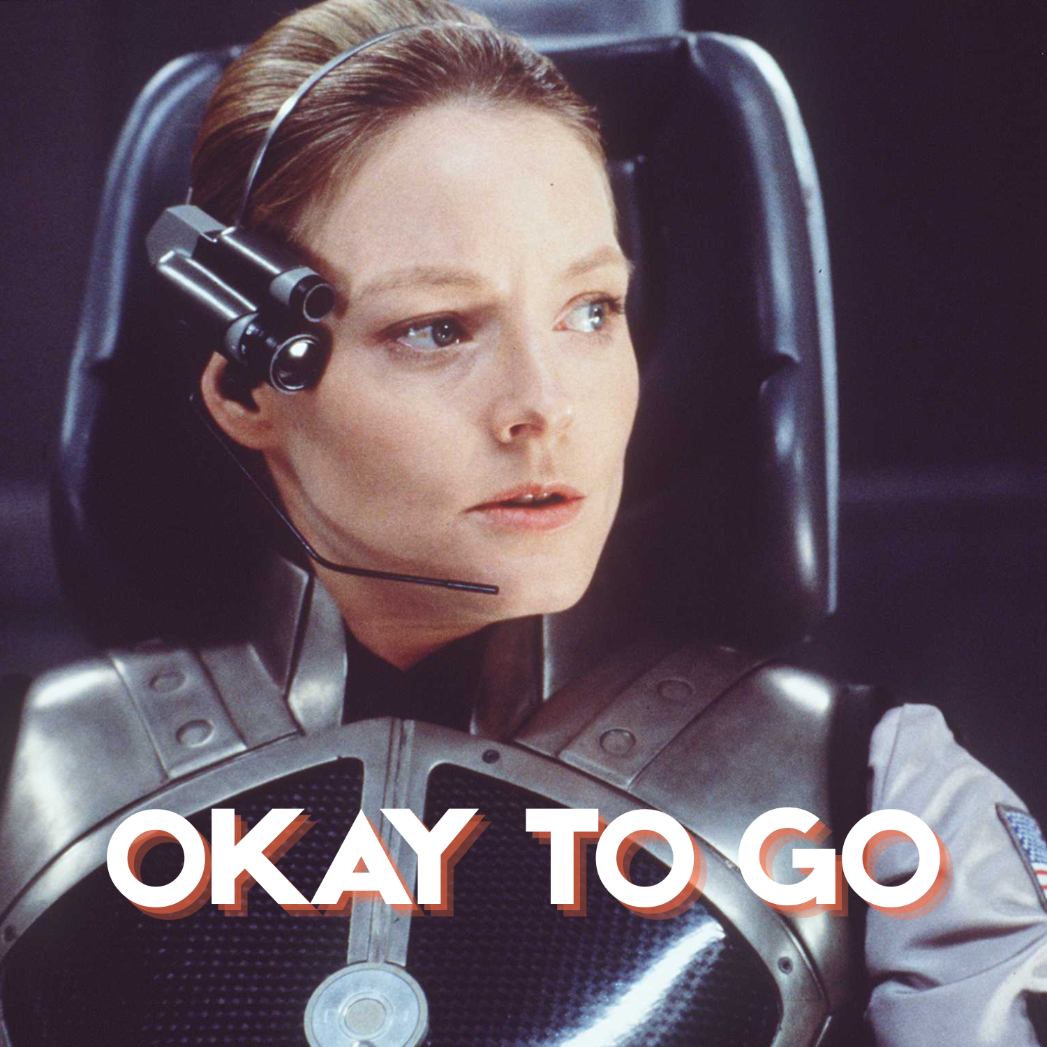 A woman with short brown hair and light skin, wearing a metallic spacesuit and a headset, sitting in a chair with a serious expression. Text overlay says 'OKAY TO GO'.