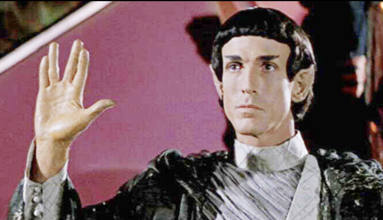 Close-up of a puppet with a human-like face, dressed in elaborate clothing, waving its hand in front of a pink background.