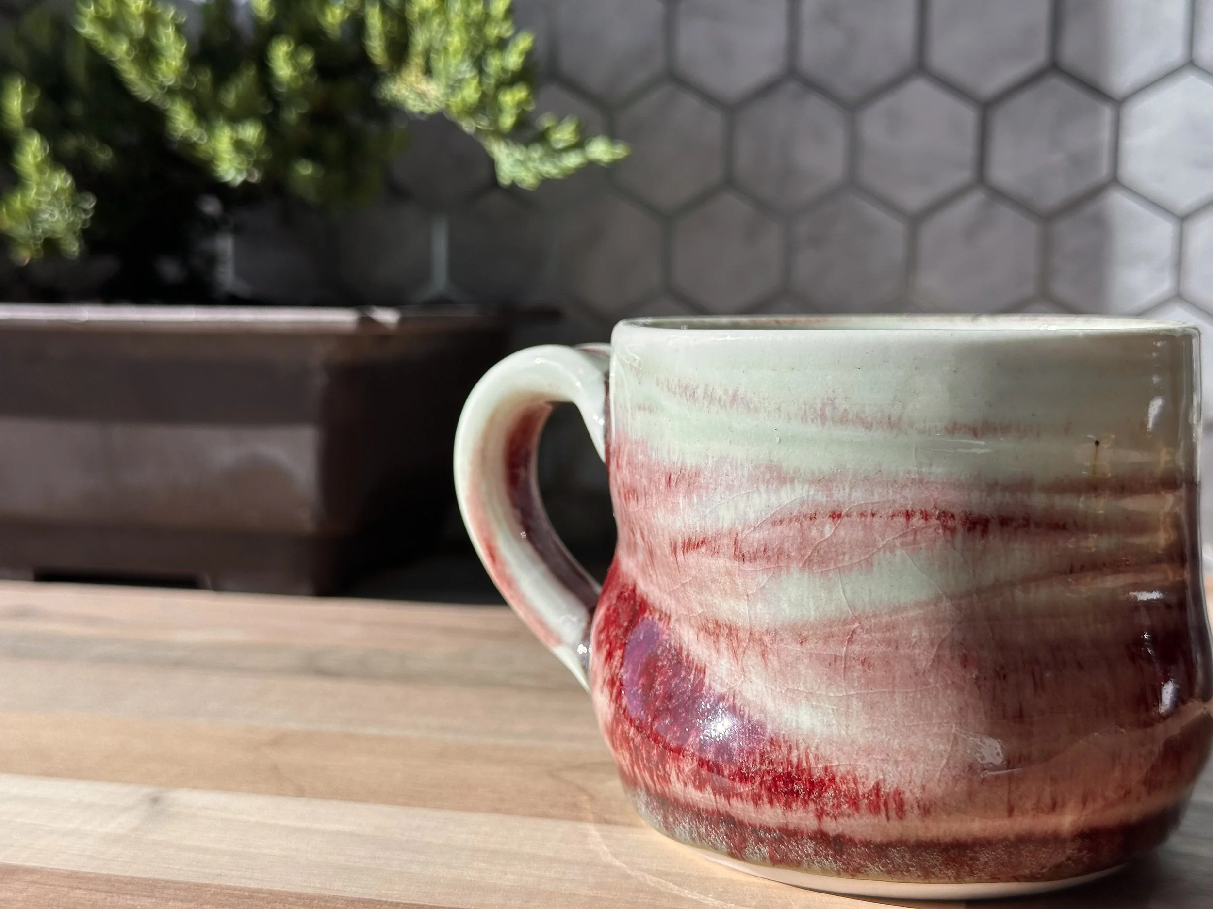 A ceramic mug with a swirl of red, pink, and white colors sitting on a wooden surface, with a plant and a hexagonal tile wall in the background.