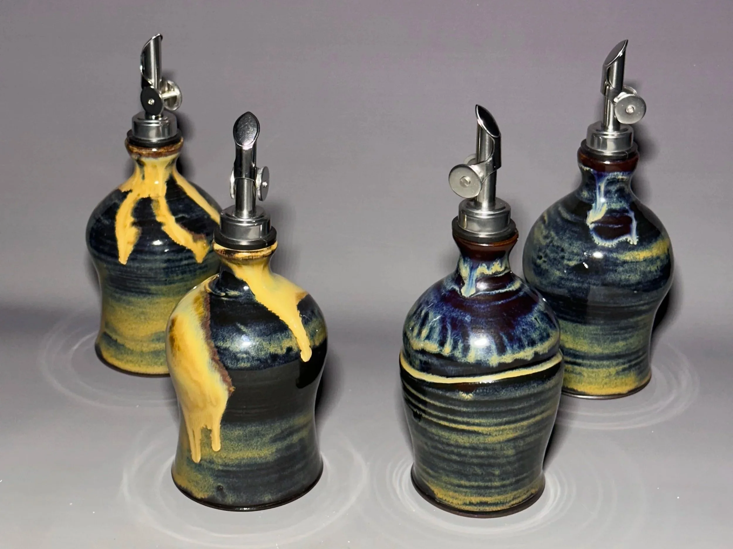 Four decorative ceramic oil bottles with metal spouts, featuring black, yellow, and blue swirl glaze patterns, placed on a gray background.