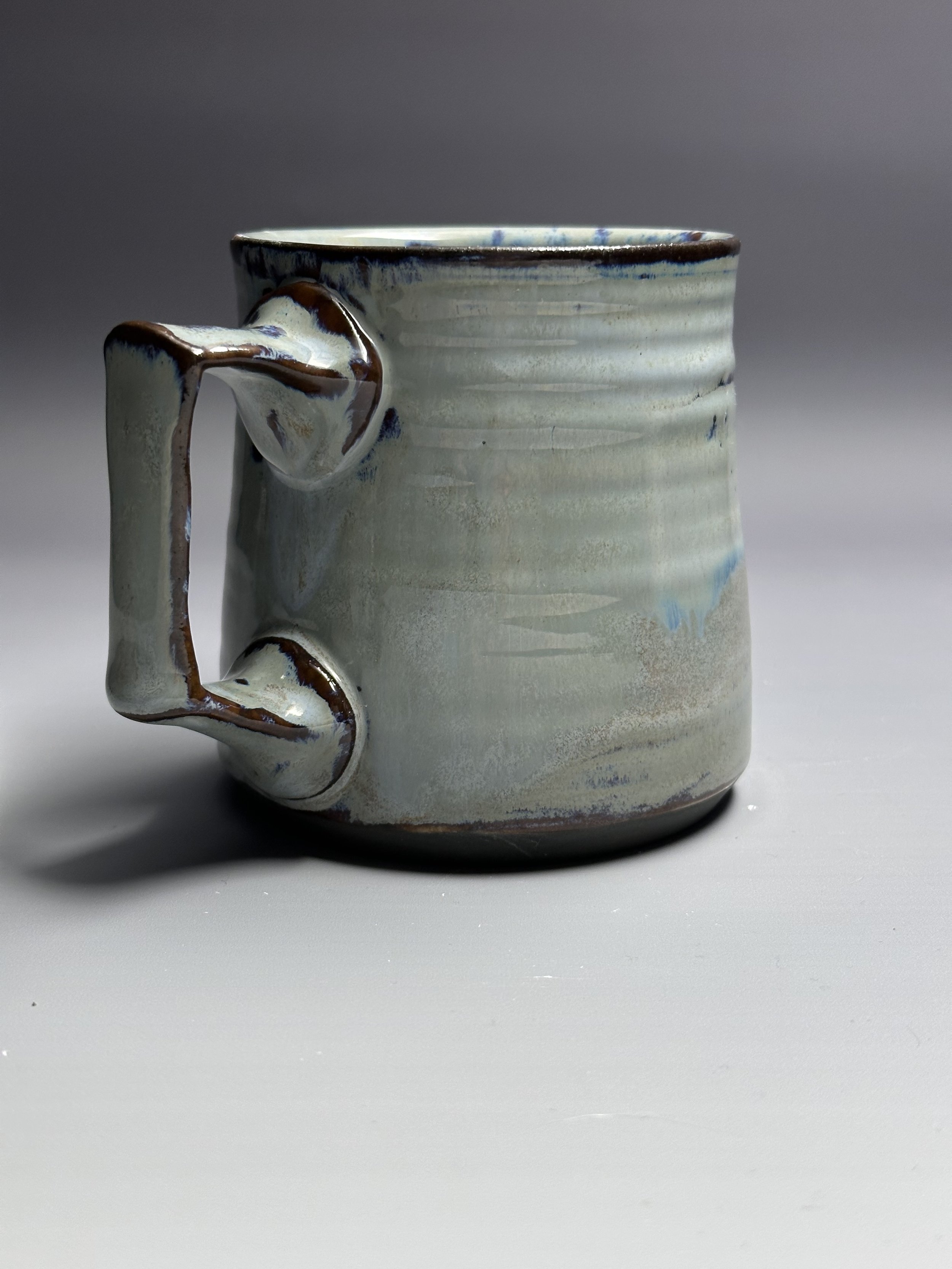 Ceramic mug with a textured off-white glaze, featuring a unique handle design with brown accents, placed on a plain light surface with a gray gradient background.