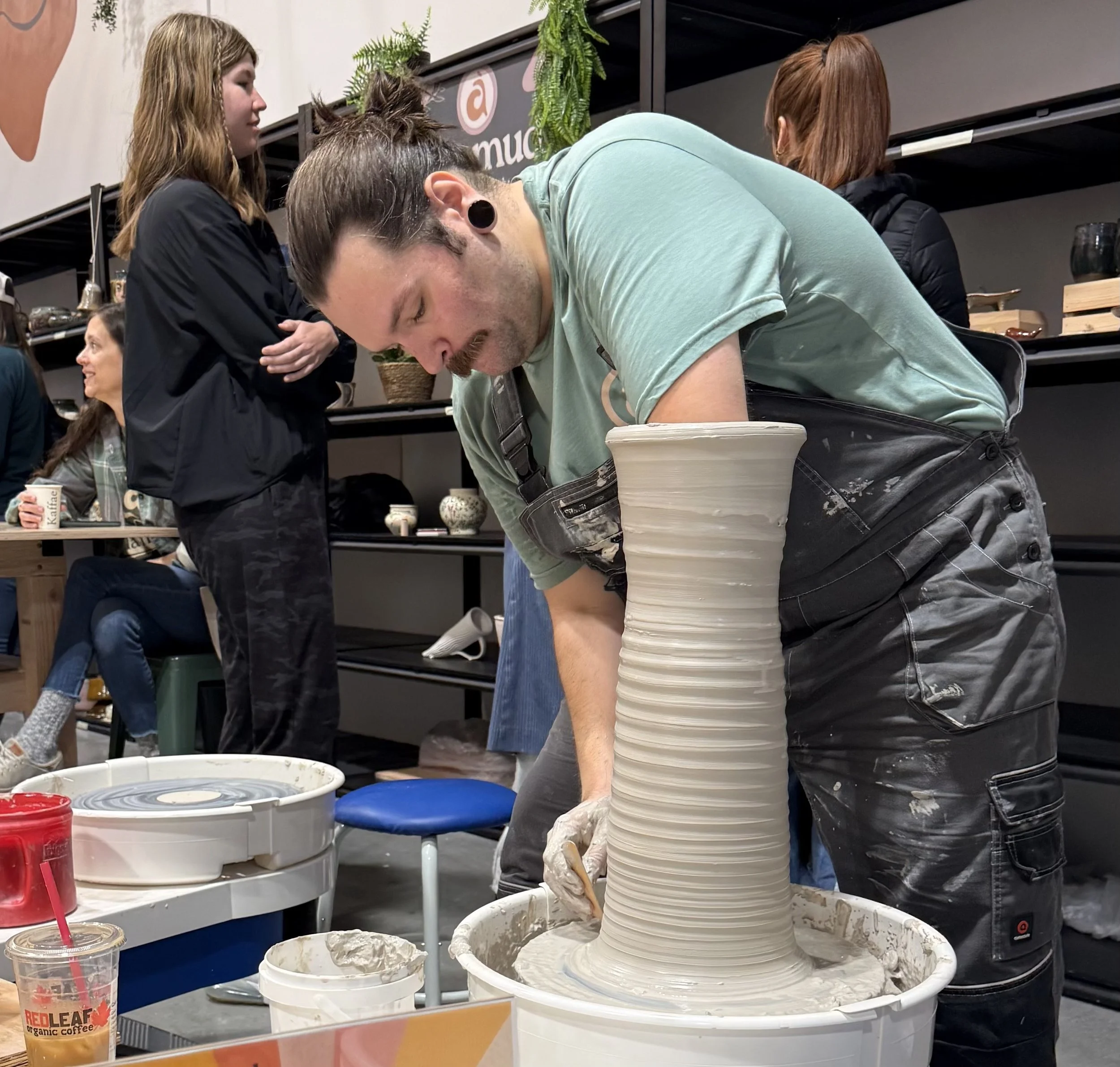 A man with gauges and a ponytail shaping a tall ceramic vase on a pottery wheel in a studio with shelves and people in the background.