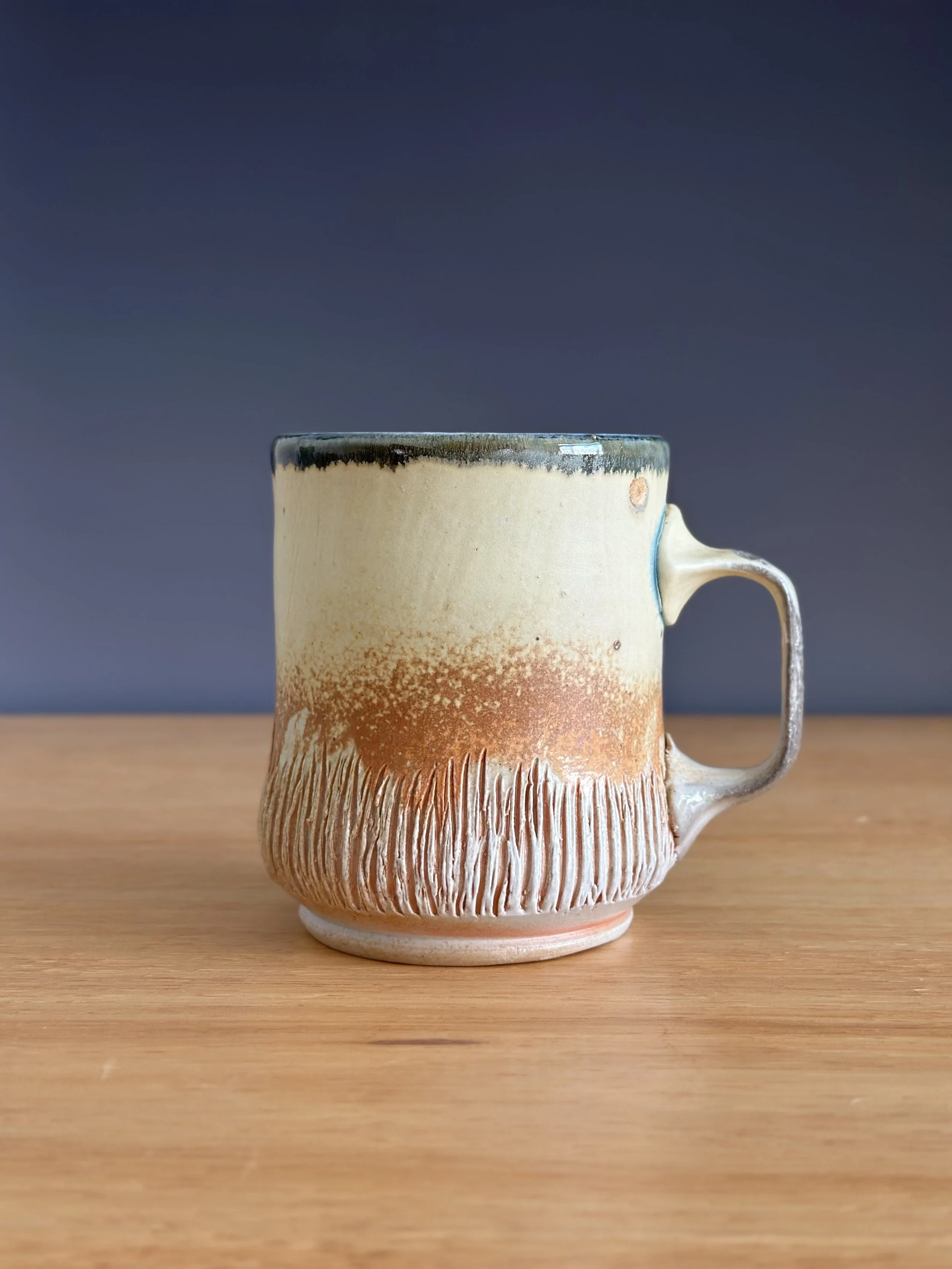 A ceramic mug with a handle, featuring a textured, earthy design with a gradient of cream, orange, and brown colors, sitting on a wooden surface against a dark grey background.