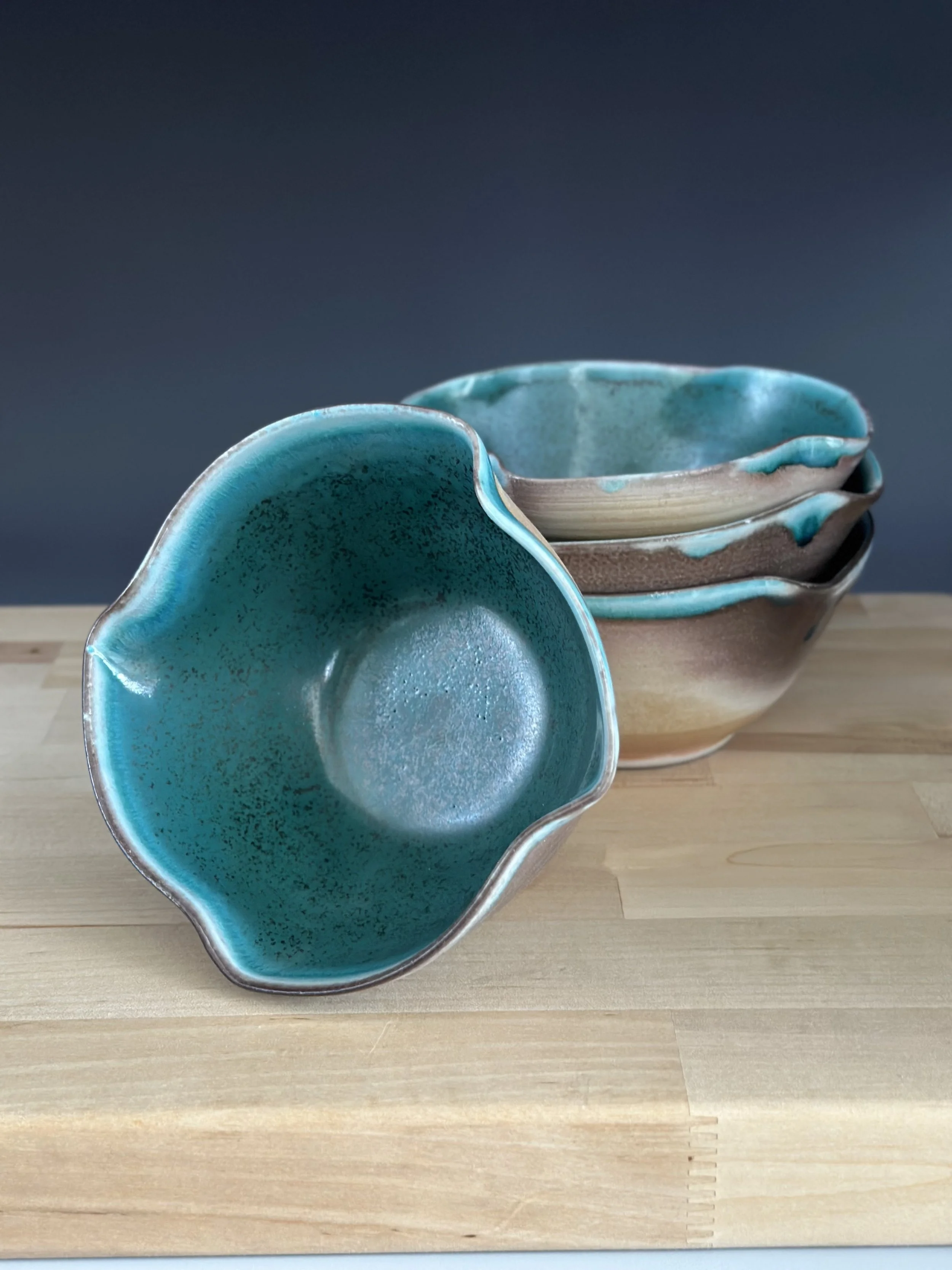 A set of five ceramic bowls with blue and earth-tone glazed finishes, stacked on a wooden surface against a gray background, with a close-up of the top bowl turned upside down.
