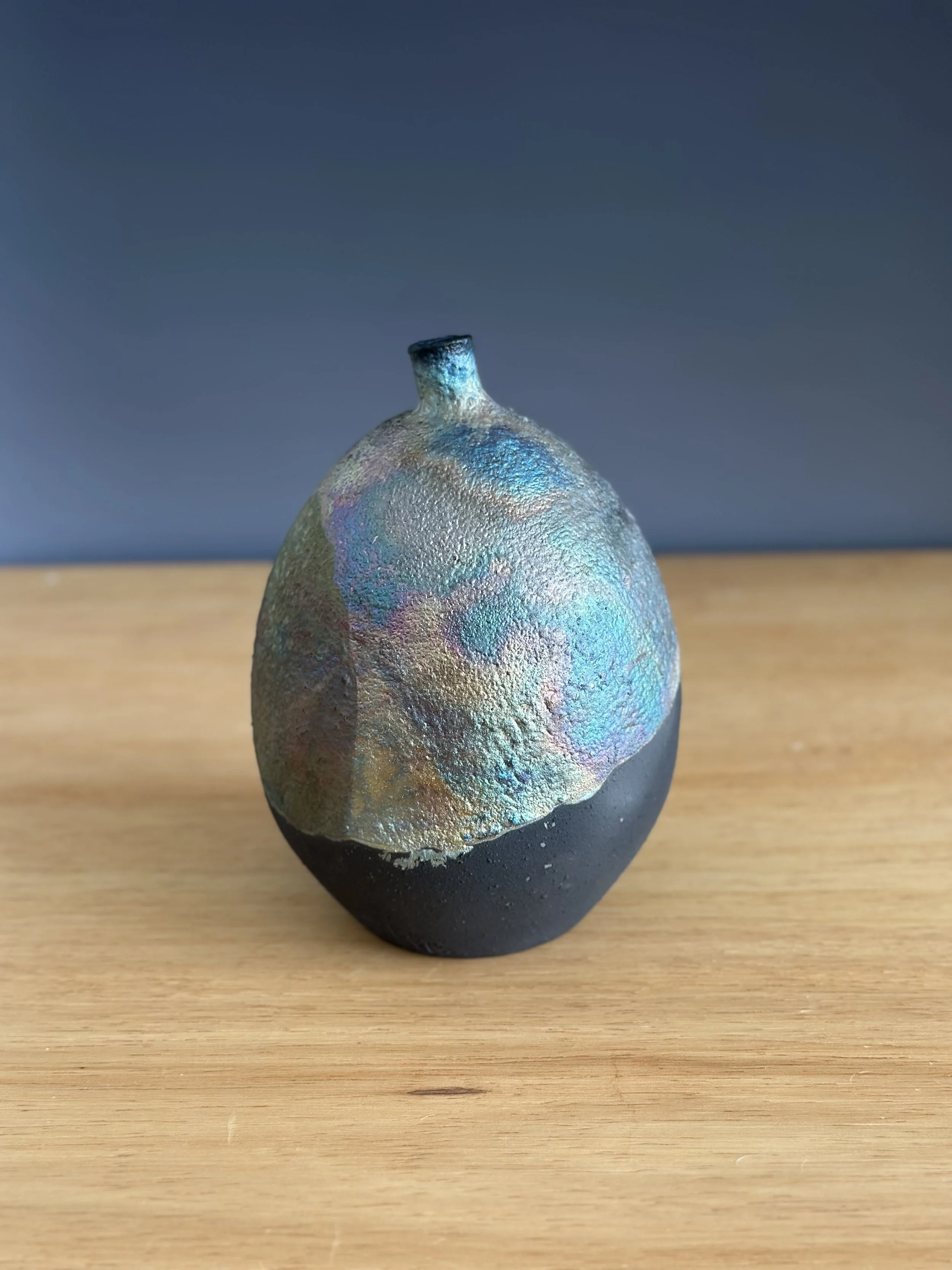 A small, round ceramic vase with a textured, iridescent upper half and a smooth, matte black lower half, placed on a wooden surface.