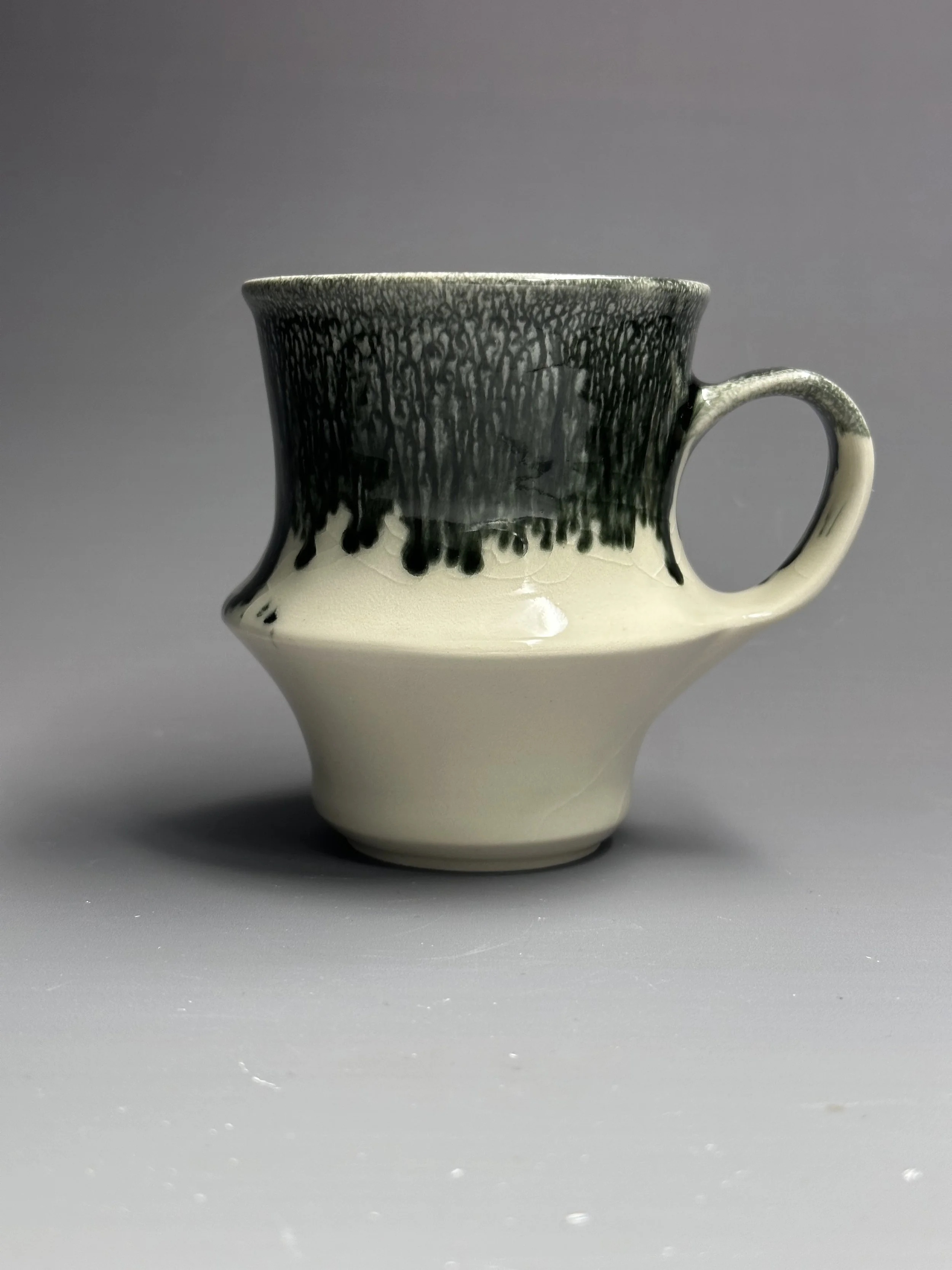 A ceramic mug with a black and cream glaze, featuring a textured dark top and smooth cream bottom, set against a gray background.