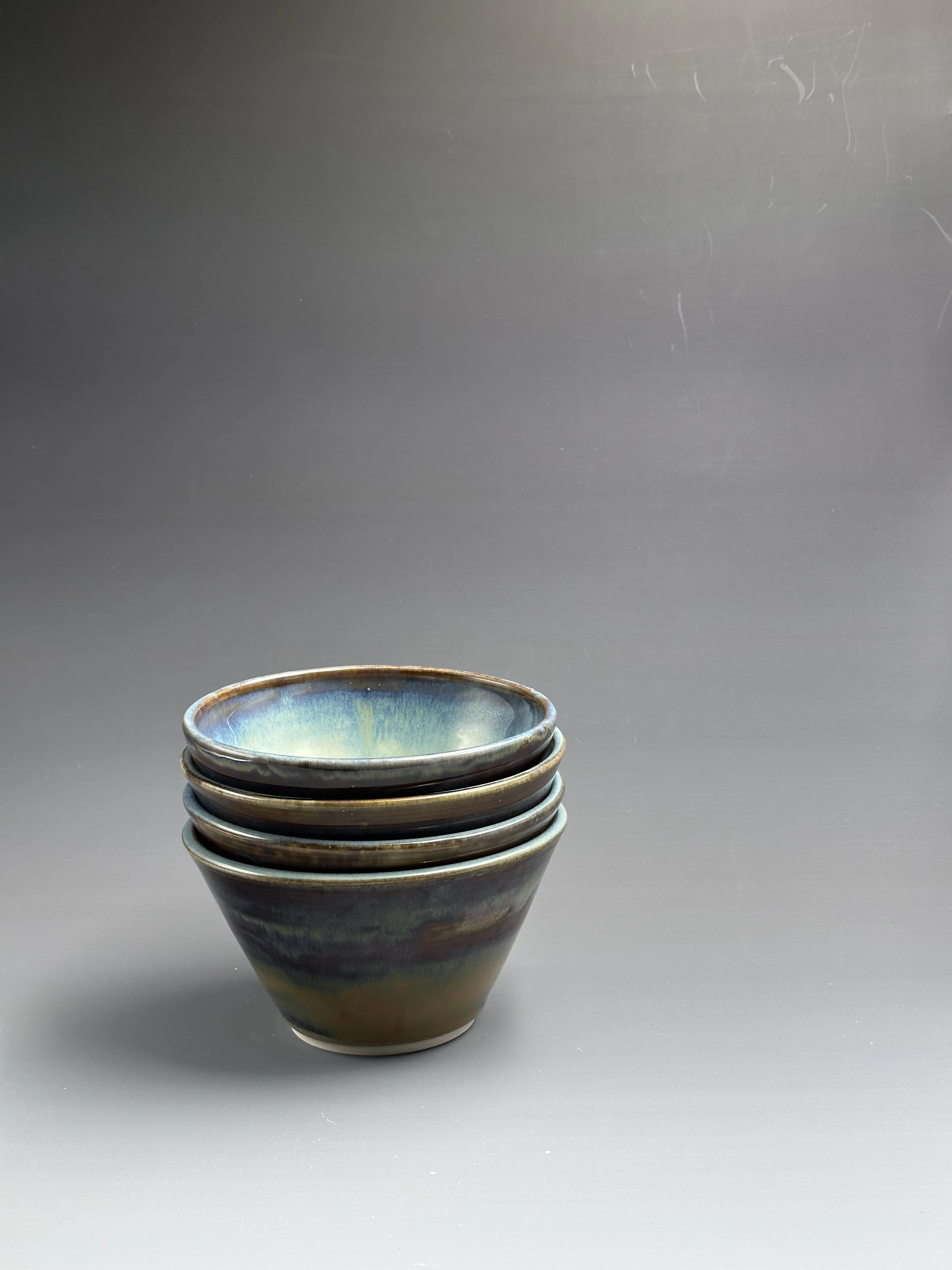A stack of five ceramic bowls with a multicolored glaze, placed on a plain gray surface against a plain gray background.