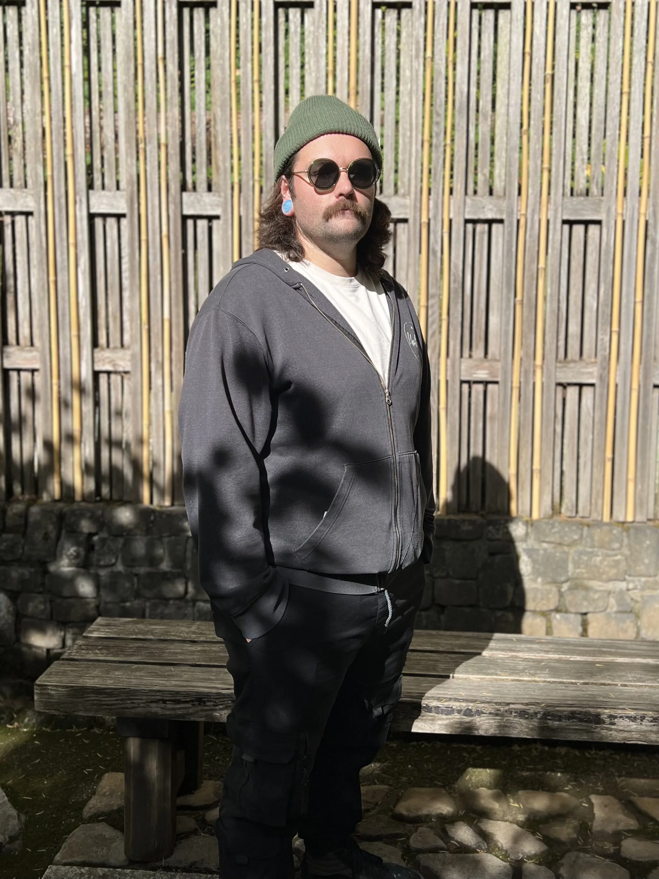 A man with long hair, a mustache, round sunglasses, a green beanie, a gray zip-up hoodie, black pants, and blue ear gauges standing outdoors in front of a wooden fence.