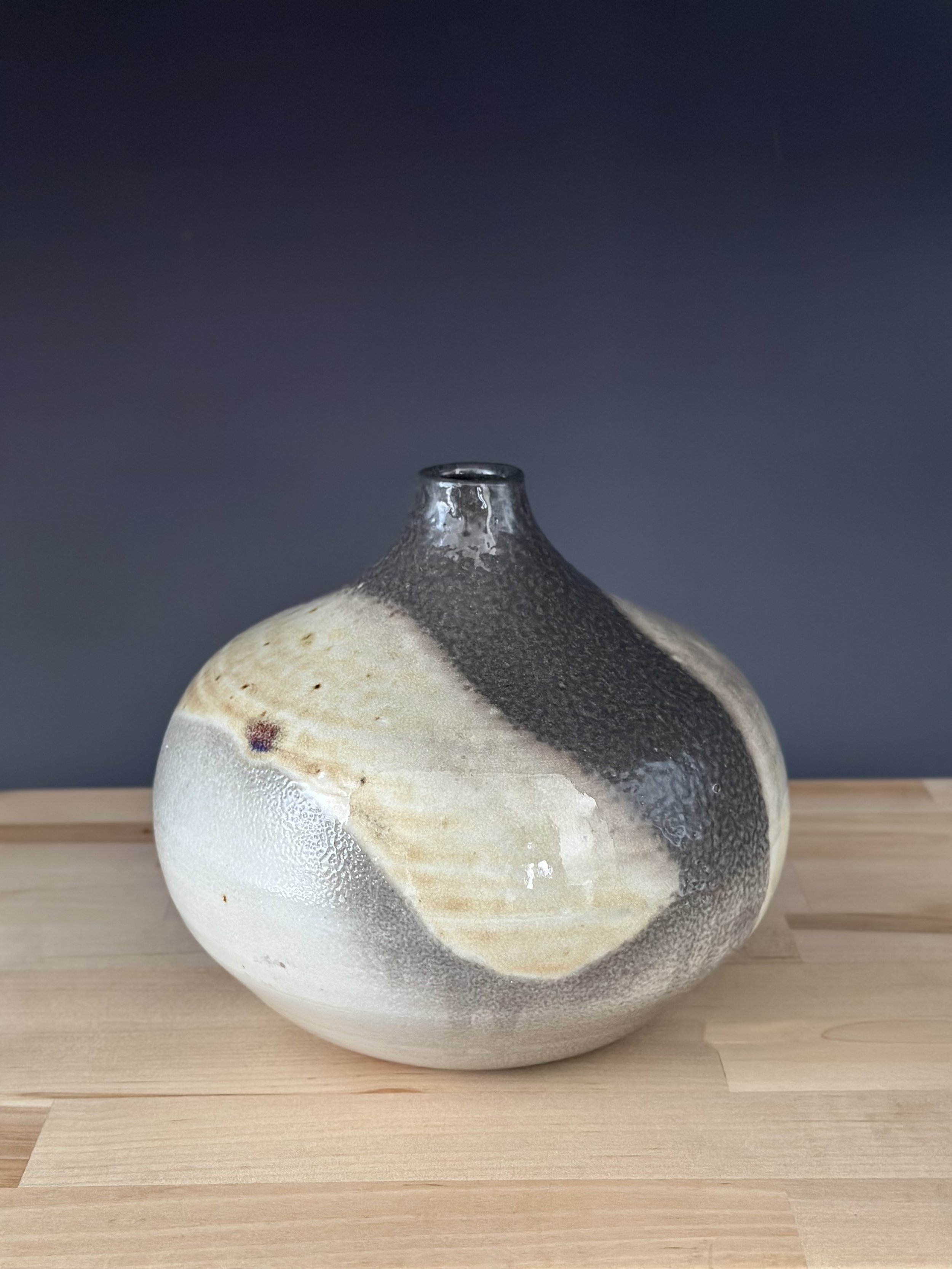 A ceramic vase with a rounded shape and narrow opening, featuring a textured, glazed surface with black, white, and beige swirl patterns, placed on a light wooden surface against a dark background.