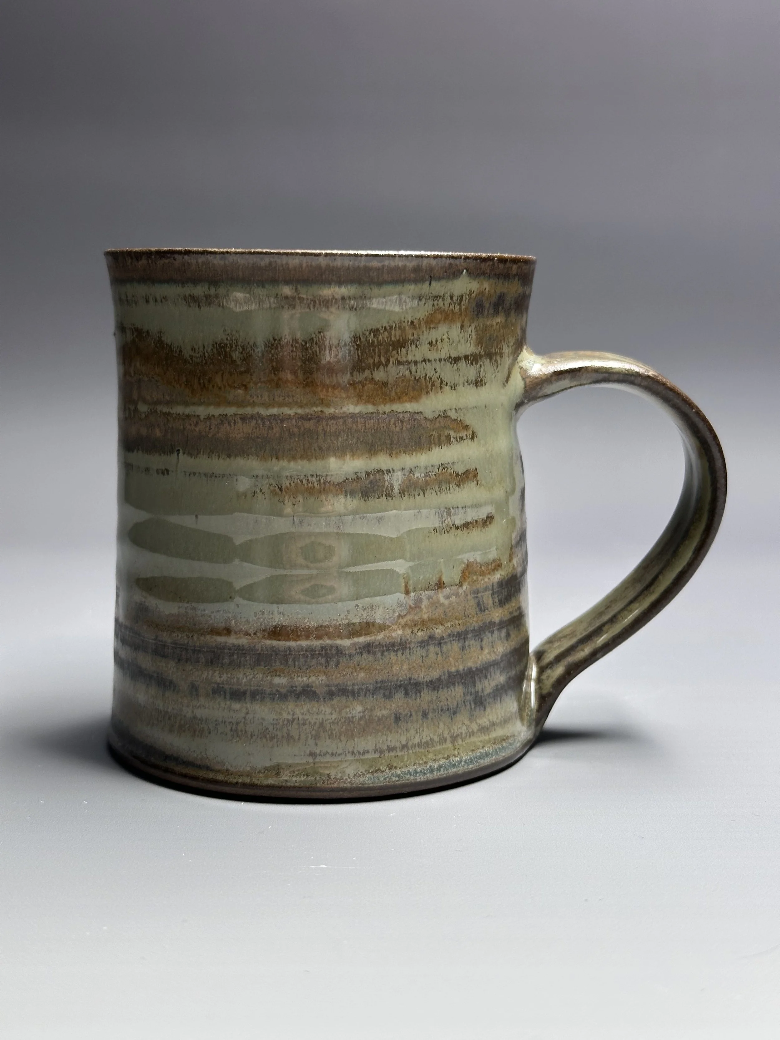 A ceramic mug with earthy, horizontal striped glaze in shades of brown, green, and beige, placed on a plain surface.