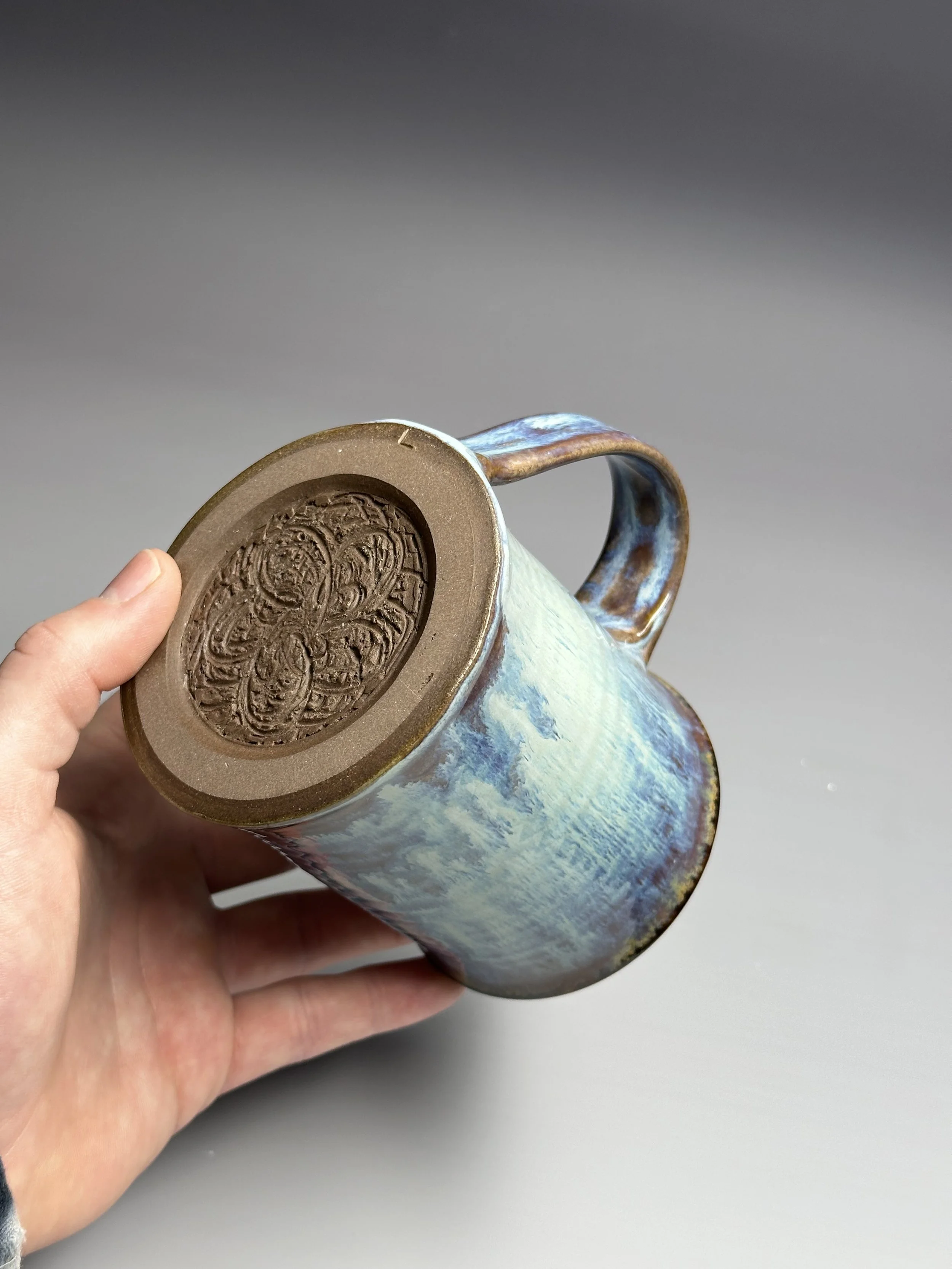 Hand holding a ceramic mug with a landscape glaze pattern and a chocolate coin on its bottom.