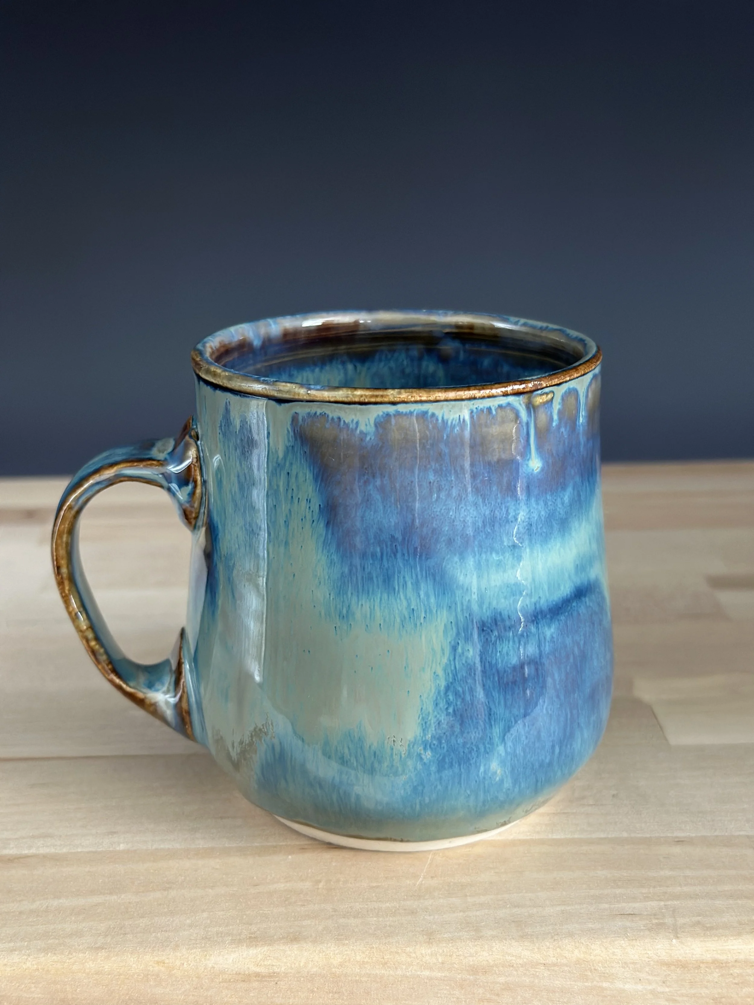 A ceramic mug with a blue, cream, and brown glaze sitting on a light wooden surface against a dark gray background.