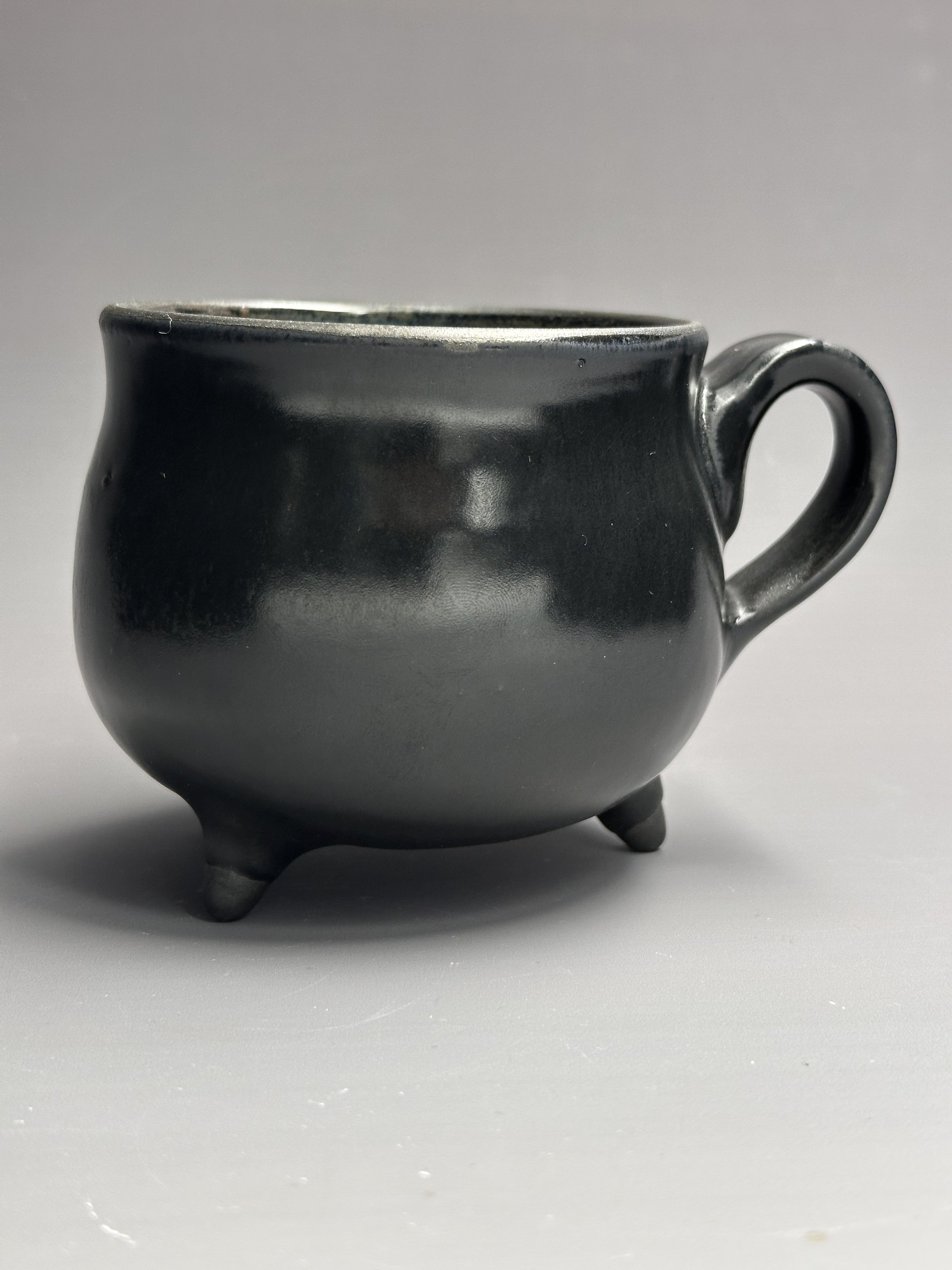 Black ceramic mug with three small legs, round body, and handle, styled with a modern, sleek design, placed on a light surface against a plain background.
