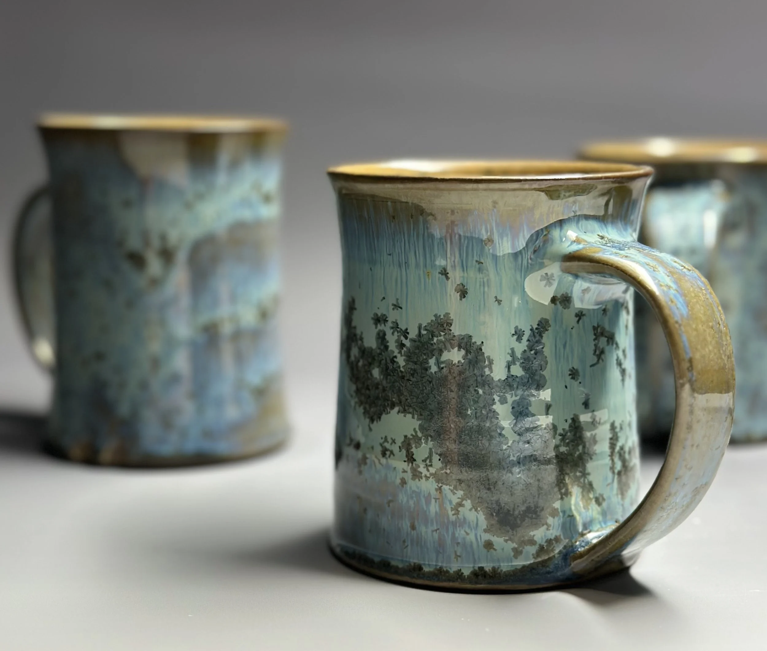 Three ceramic mugs with a blue and gold glaze finish, arranged on a white surface with a gray background.