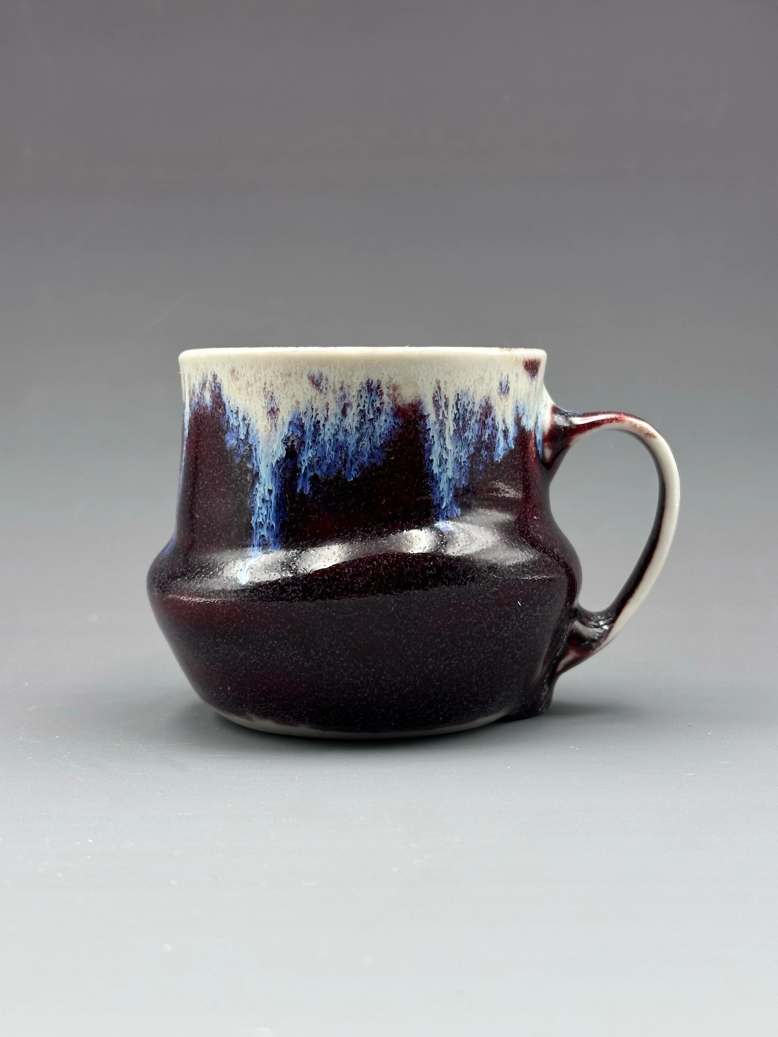 A ceramic mug with a dark purple and blue glaze, featuring an abstract drip pattern near the rim, set against a plain gray background.