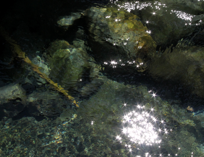 Clear water with rocks and stones beneath the surface, reflecting sunlight creating sparkling effects.