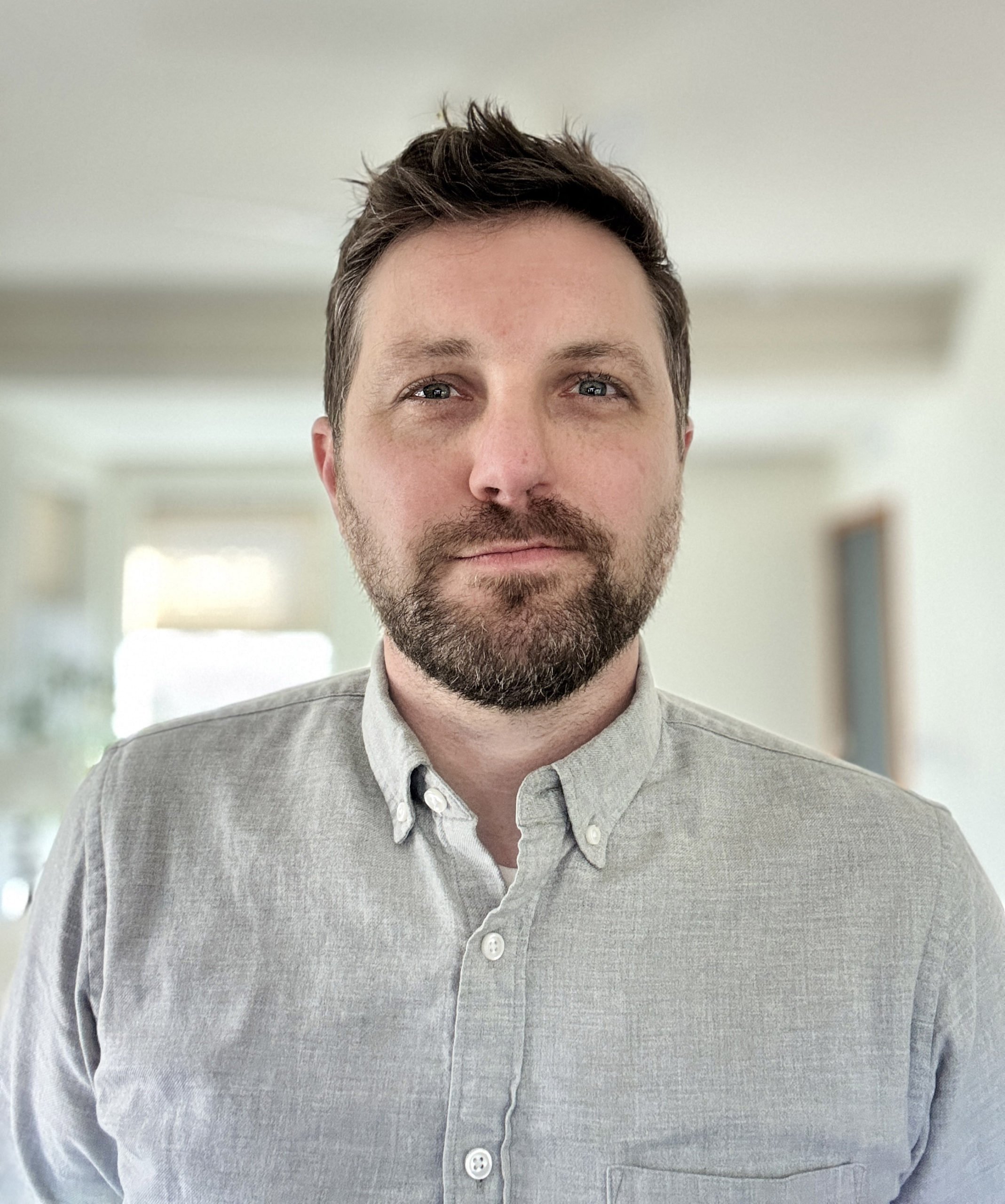 A man with brown hair and a beard wearing a light gray button-down shirt, standing indoors with a blurred background.