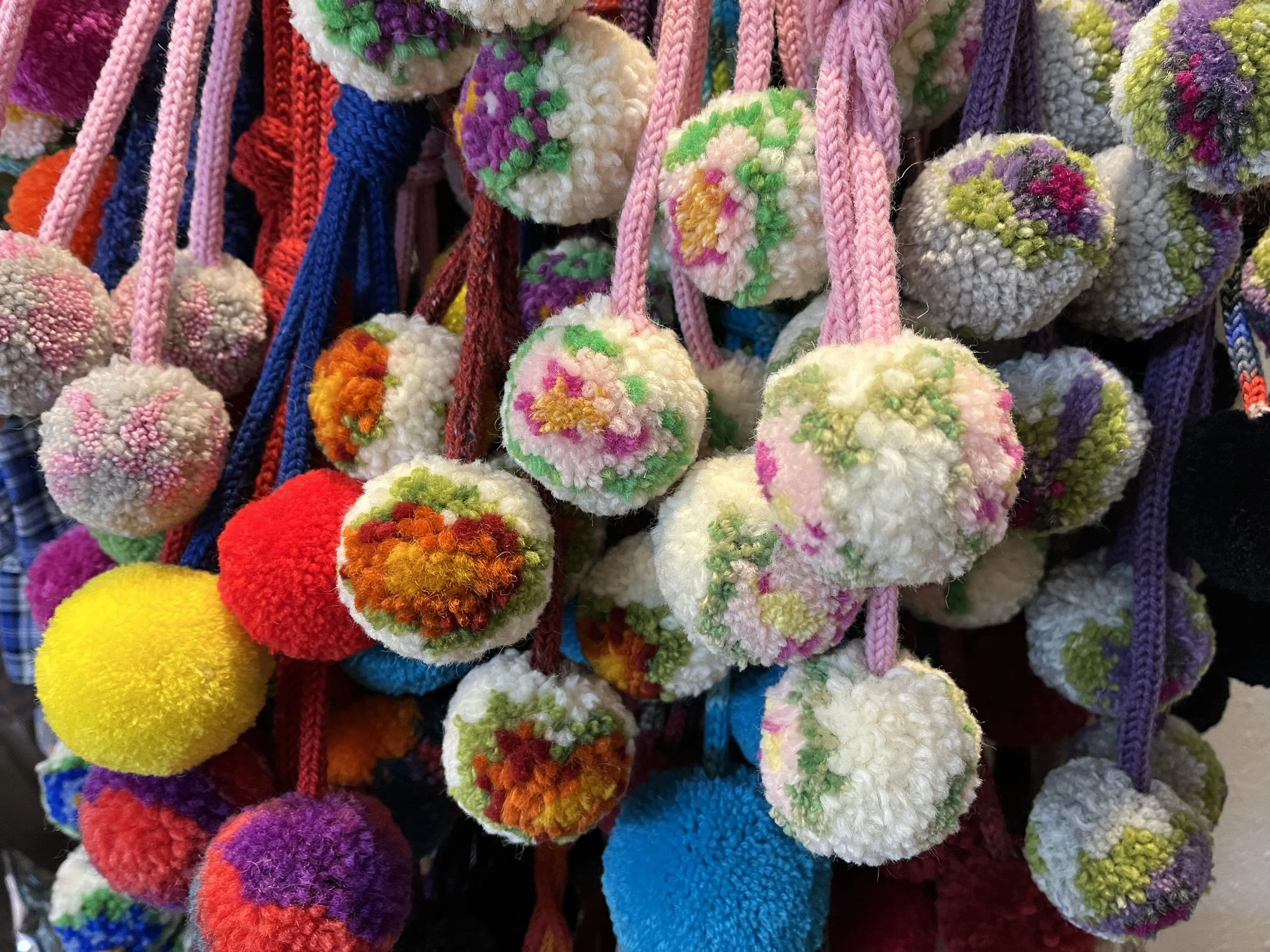 A close-up of colorful pom-pom keychains made of fuzzy material with floral patterns, hanging on pink, purple, red, and blue cords.