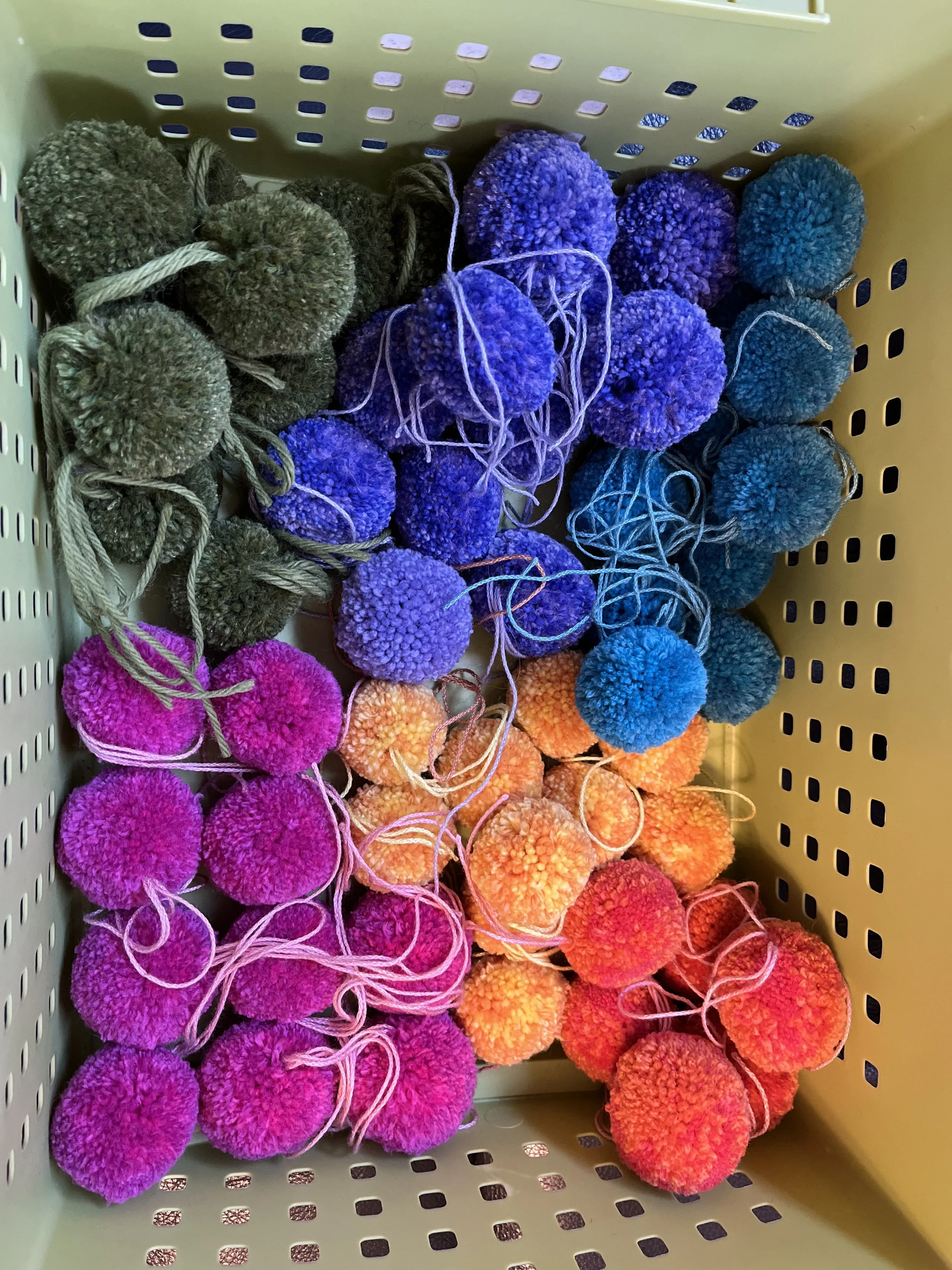 A collection of colorful yarn pom-poms in green, purple, blue, pink, orange, and red stored in a plastic basket.