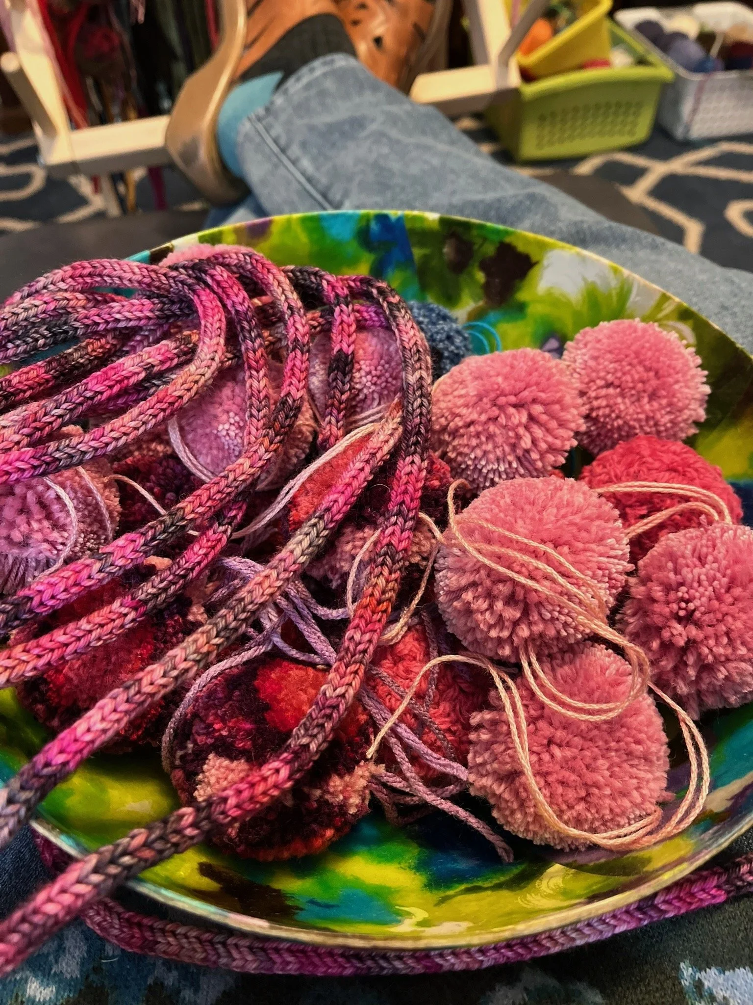 A colorful bowl filled with pink pom-poms and yarn, with a person sitting back with legs crossed, wearing blue jeans and brown shoes in the background.