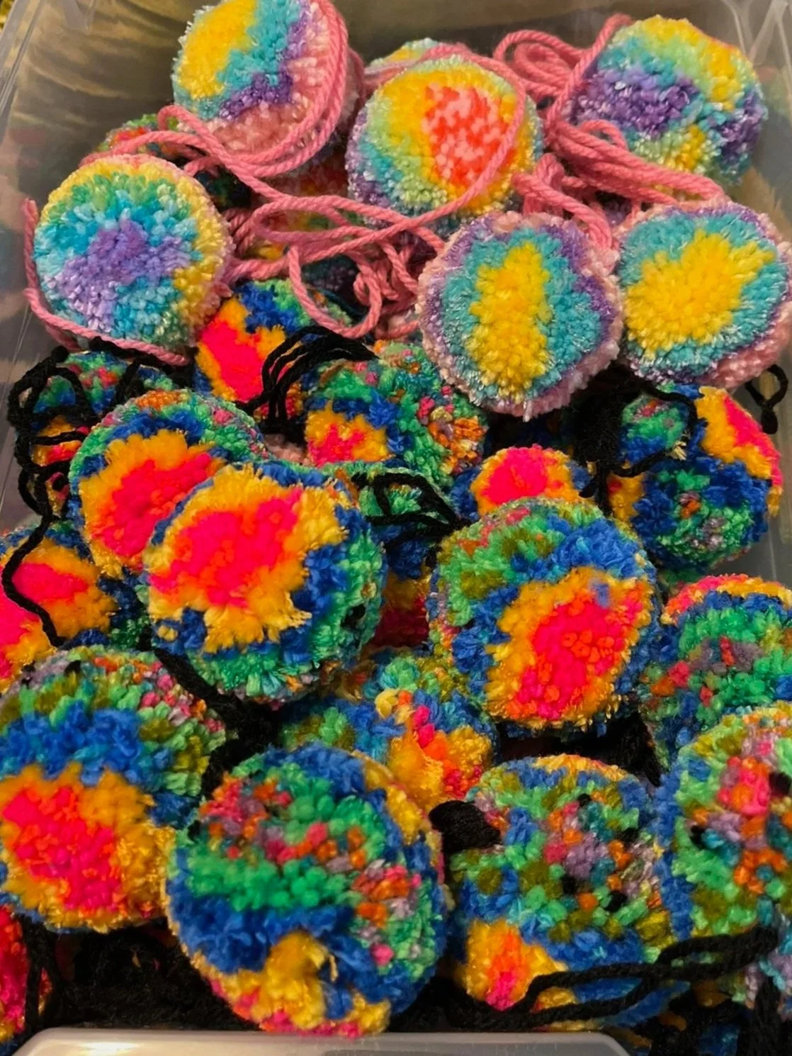 Colorful handmade pom-pom keychains with rainbow and floral patterns in a plastic container.