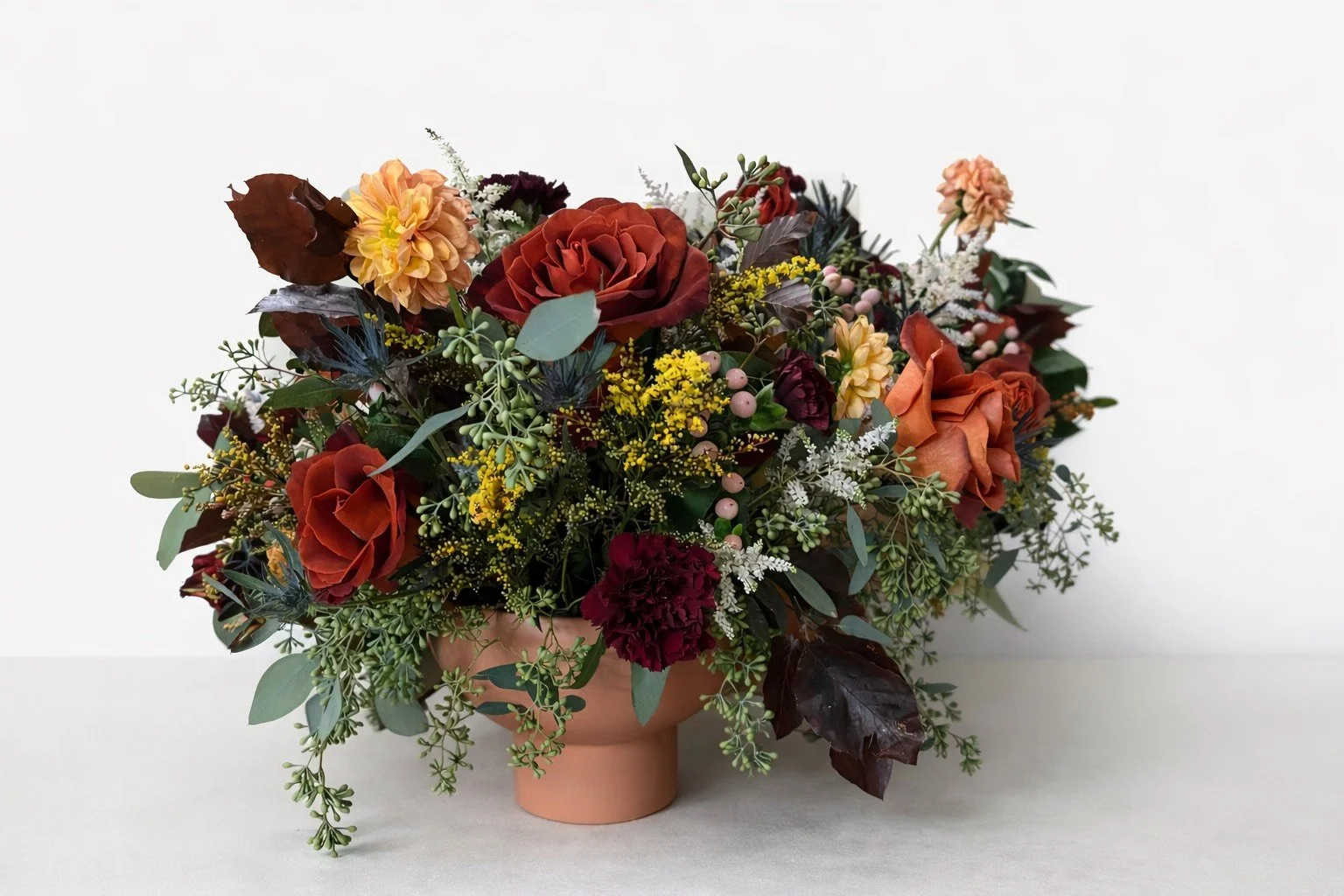 A floral arrangement in a peach-colored vase with red, orange, yellow, and burgundy flowers, surrounded by greenery and small decorative berries, set against a plain white background.