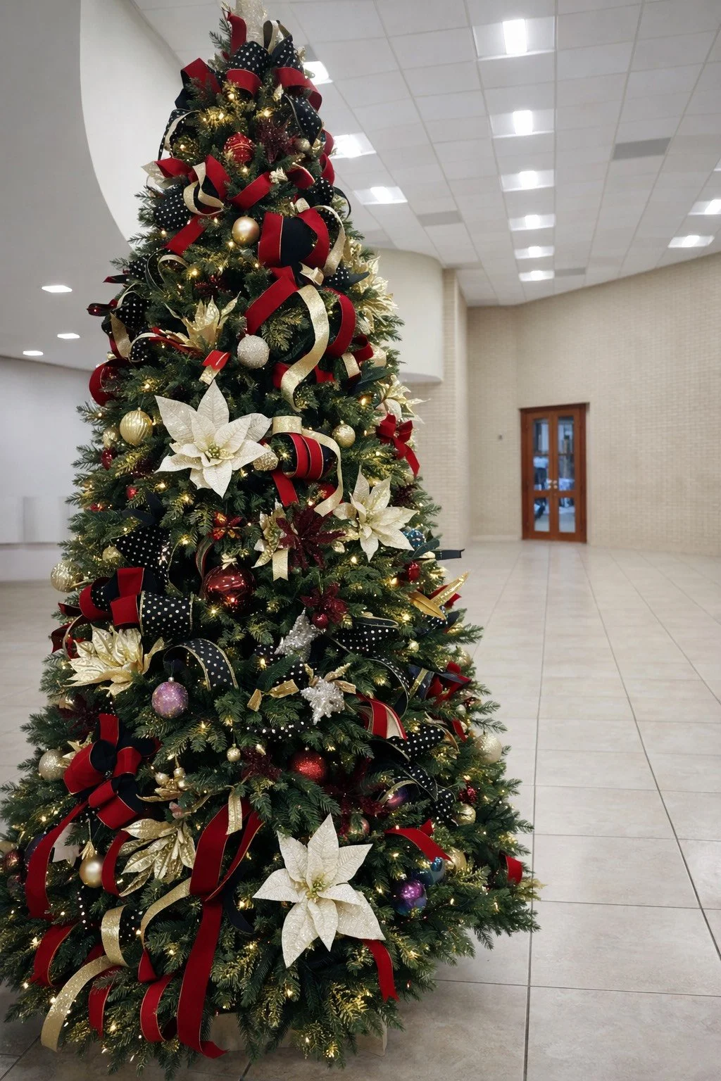 A decorated Christmas tree with red, black, and gold ribbons, white poinsettia flowers, and gold and purple ornaments, located in a spacious, well-lit indoor area.
