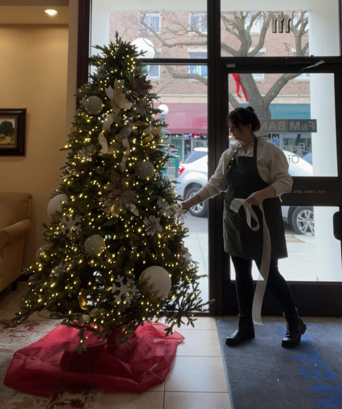 A woman decorating a Christmas tree with white and gold ornaments and lights inside a building.
