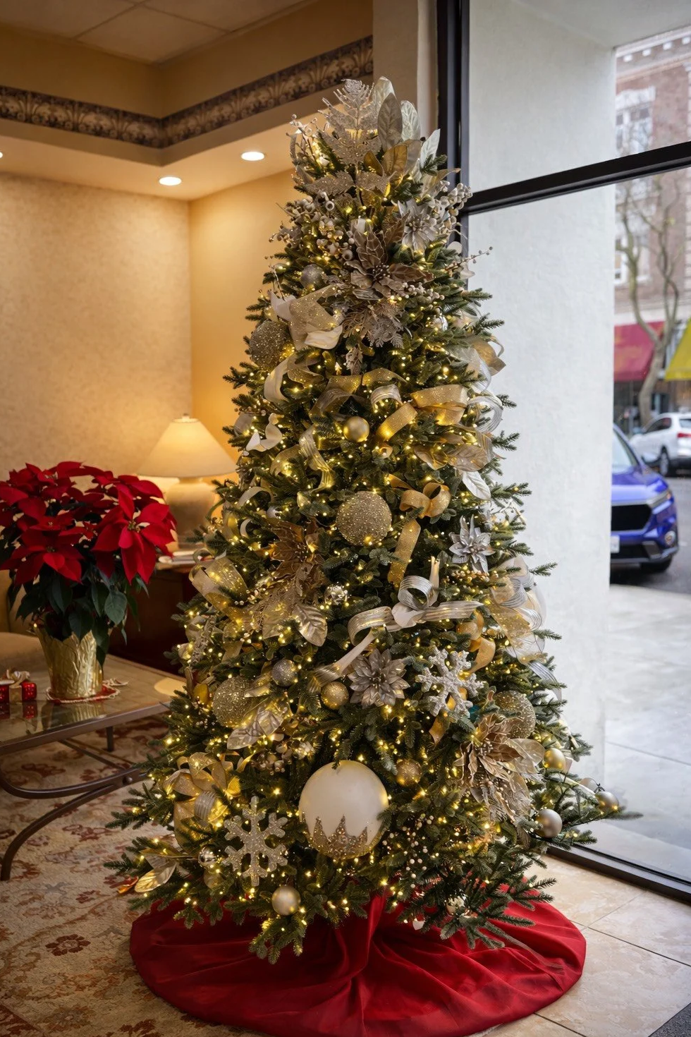 Decorated Christmas tree with gold, silver, and white ornaments and ribbons, illuminated with string lights, placed near a window in a festive living room.