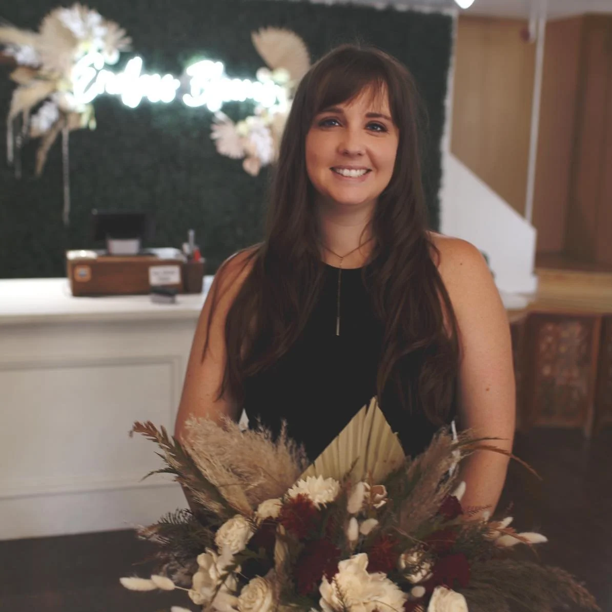 A woman with long brown hair and a black dress smiling while holding a bouquet of dried flowers indoors. In the background, there is a neon sign with white lettering that says 'Gather Together,' decorated with flowers and pampas grass.