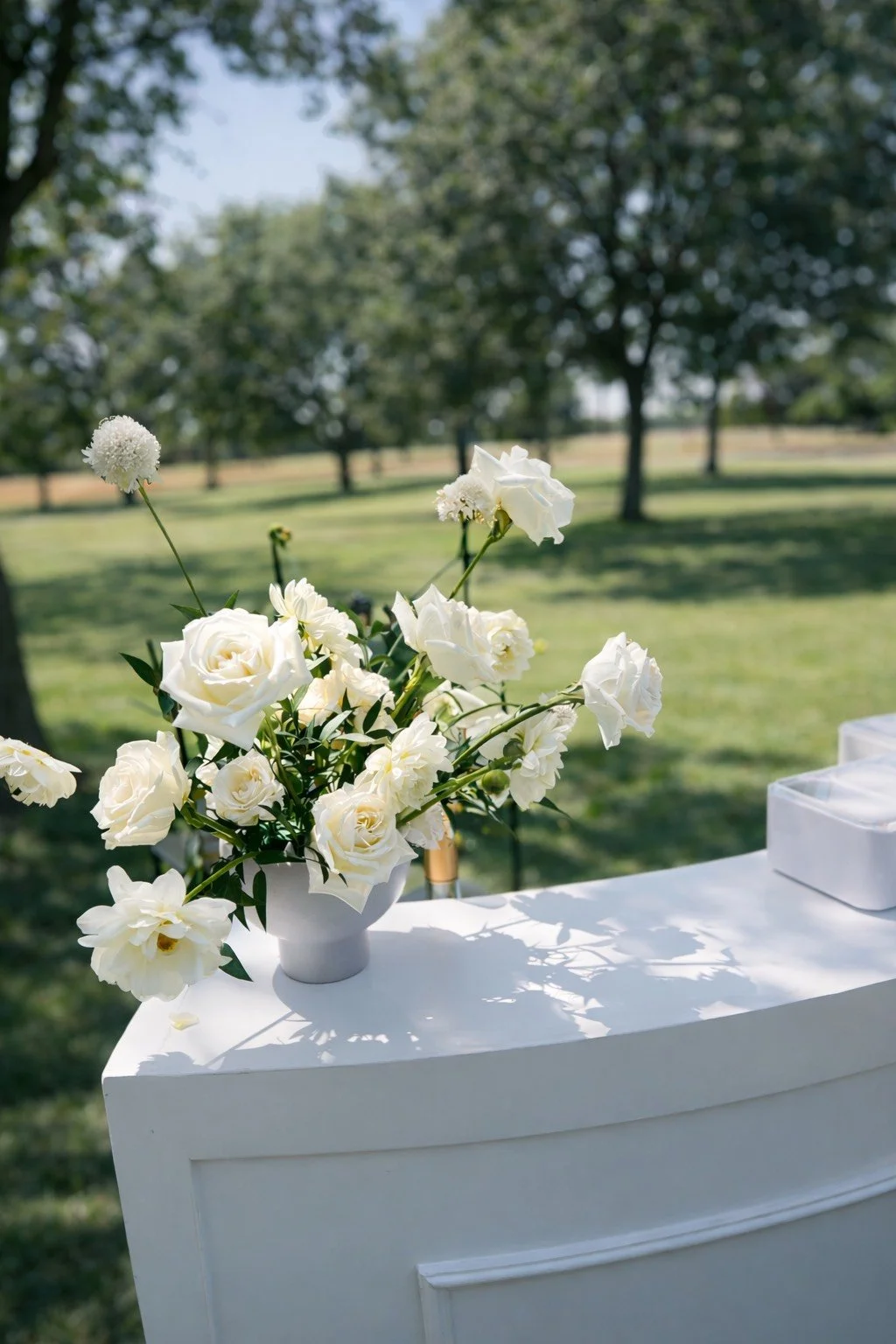 A white floral arrangement with roses, peonies, and other flowers in a white vase on a white table outdoors, with green trees and grass in the background.
