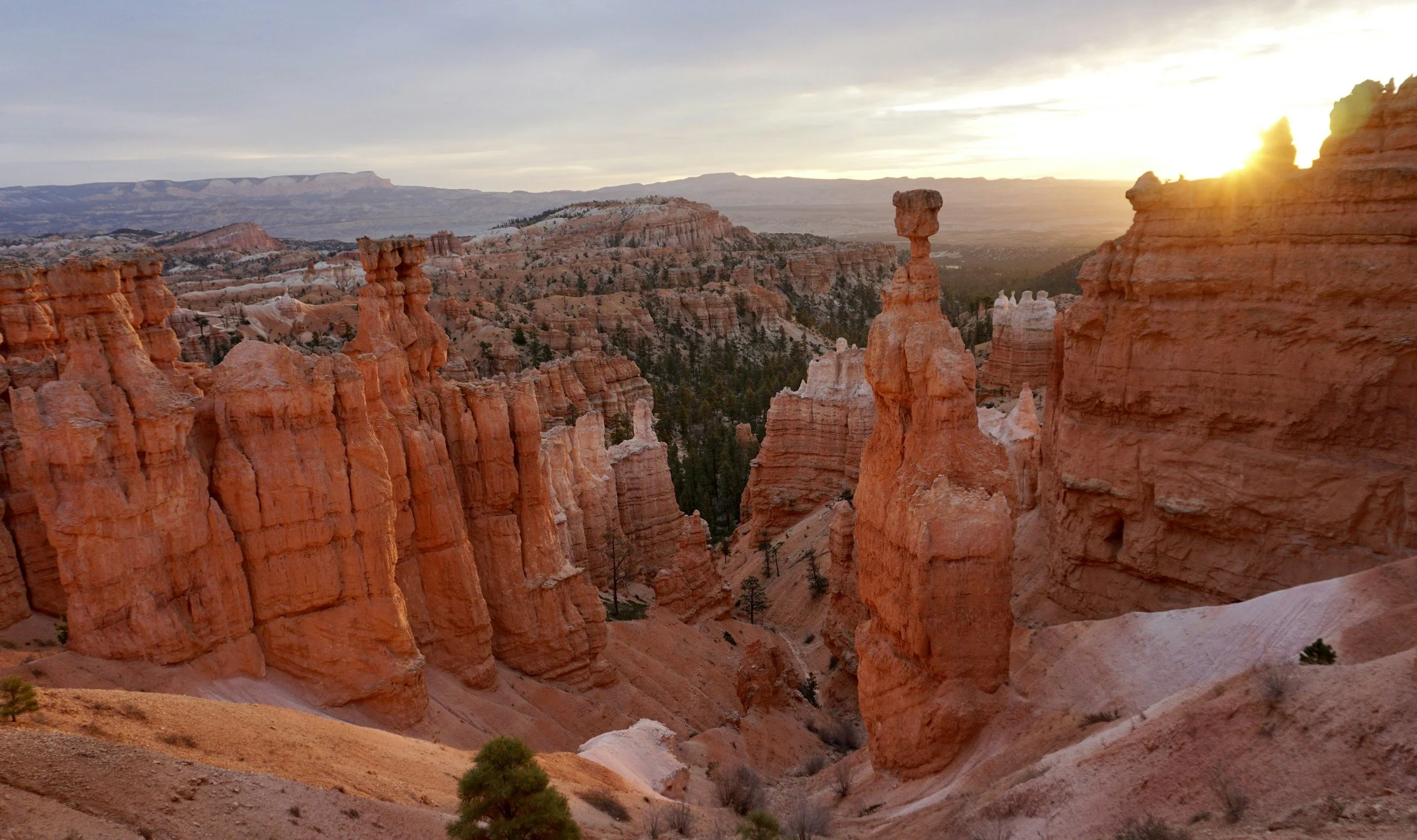 Sunset over the hoodoos and rock formations in Bryce Canyon National Park, with a cloudy sky and distant mountains.