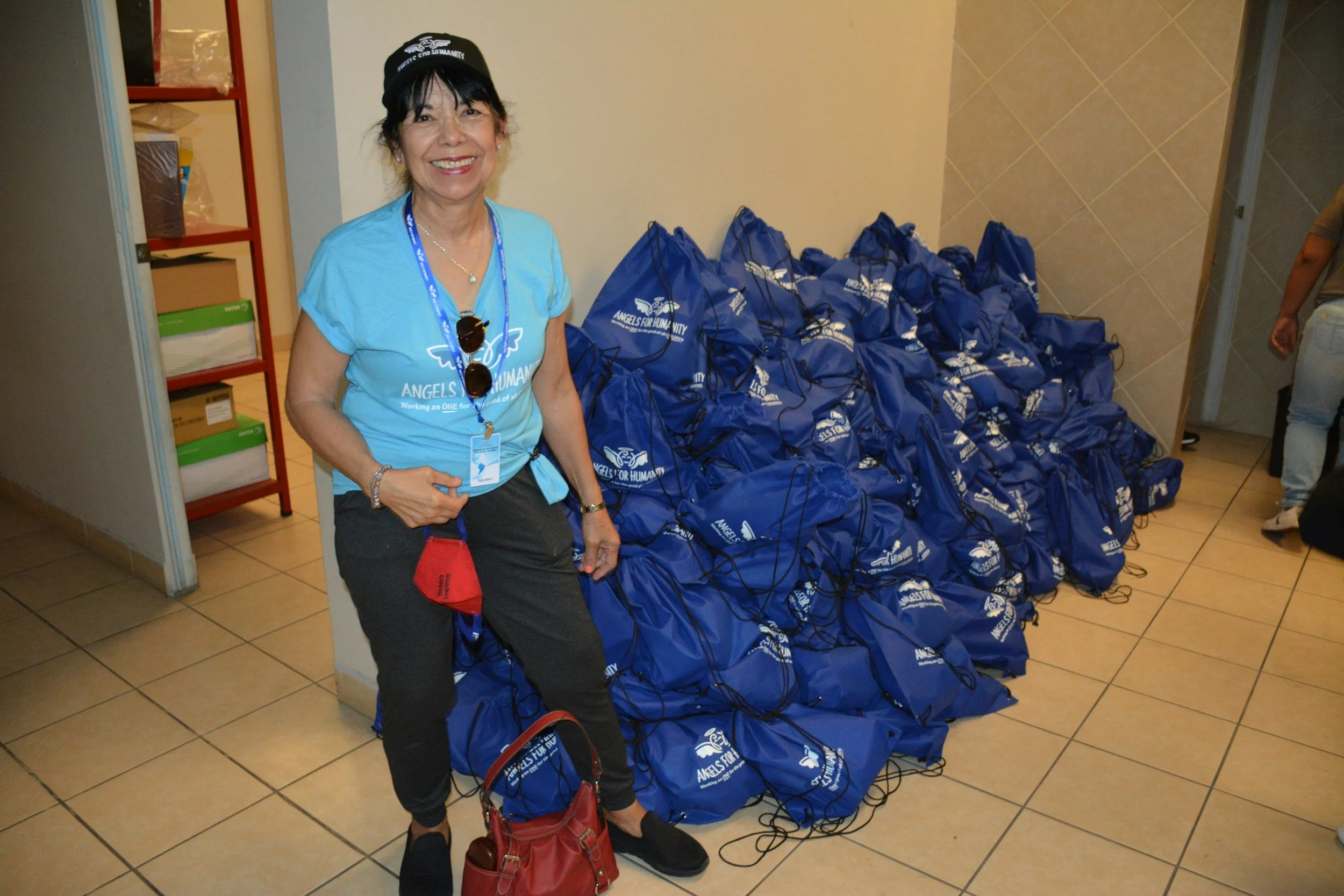 A woman smiling standing beside a large pile of blue drawstring bags with white logos, happy that she is helping with donations for a nonprofit serving survivors of domestic violence and sexual assault.