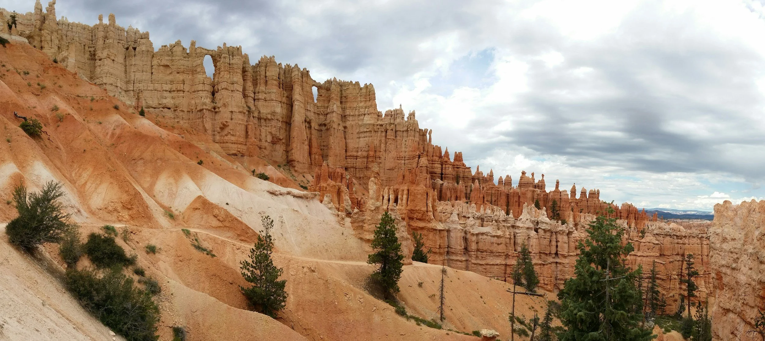 View of colorful hoodoos and cliffs in Bryce Canyon National Park, with a cloudy sky overhead and sparse trees on the landscape.