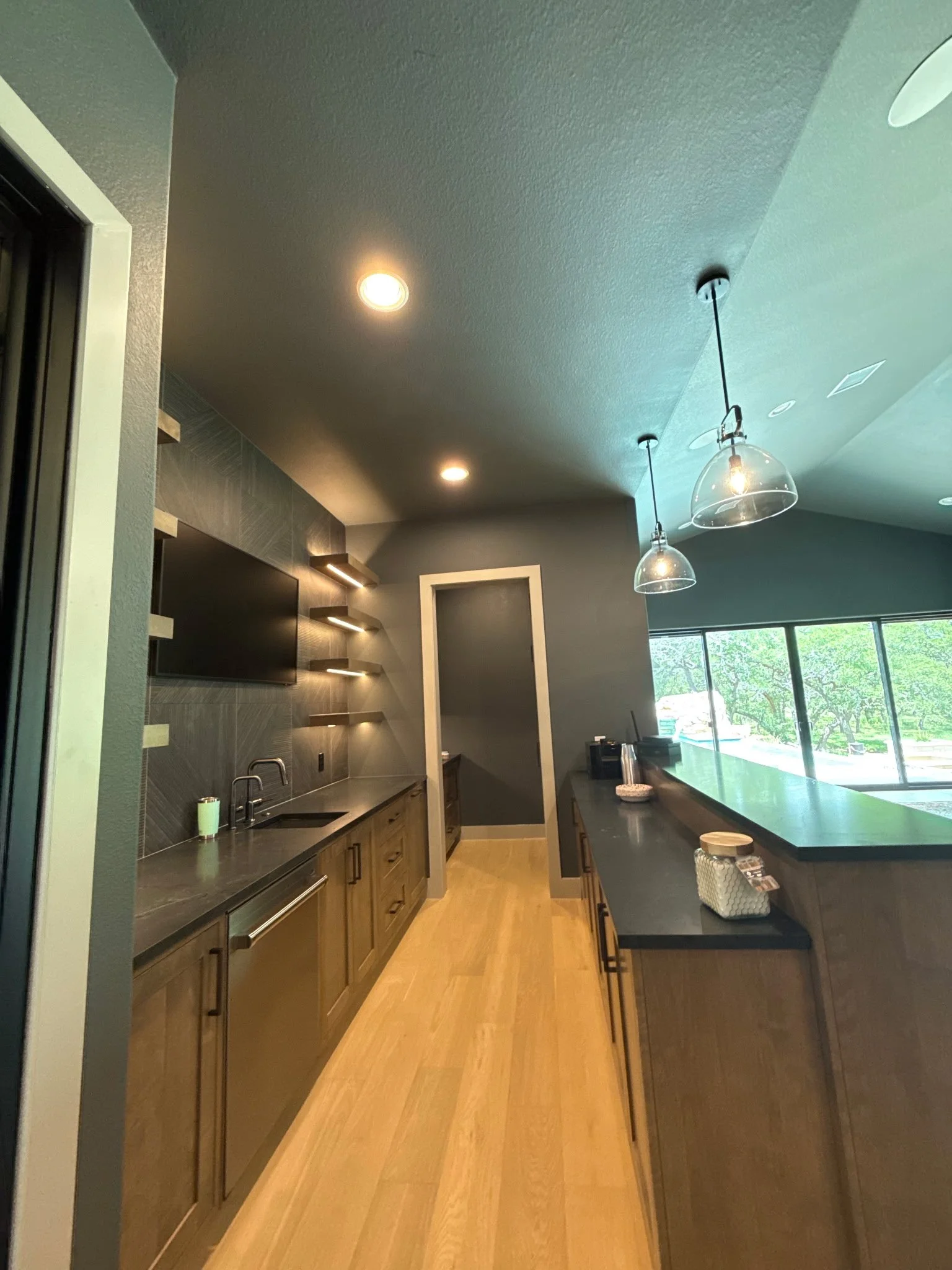 Modern kitchen with dark cabinets, a black countertop, a wall-mounted TV, open shelves, and large windows with a view of trees outside.